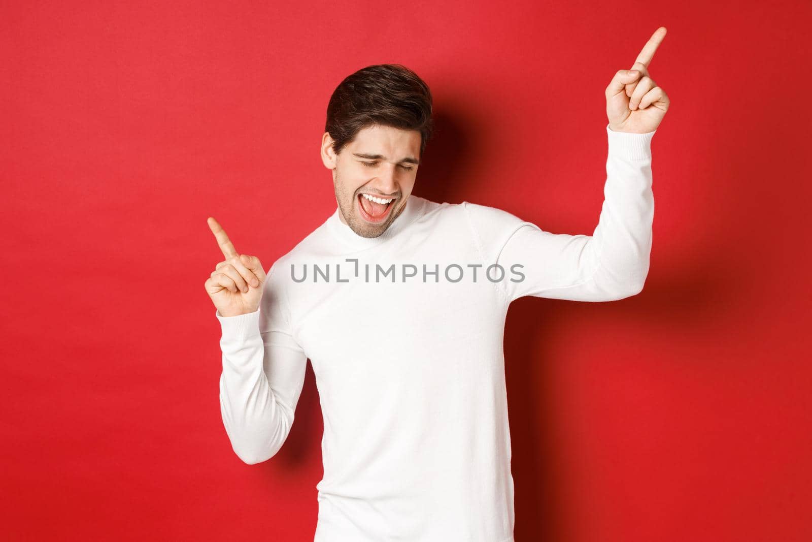 Portrait of handsome man enjoying new year party, dancing and having fun, pointing fingers up, standing over red background.