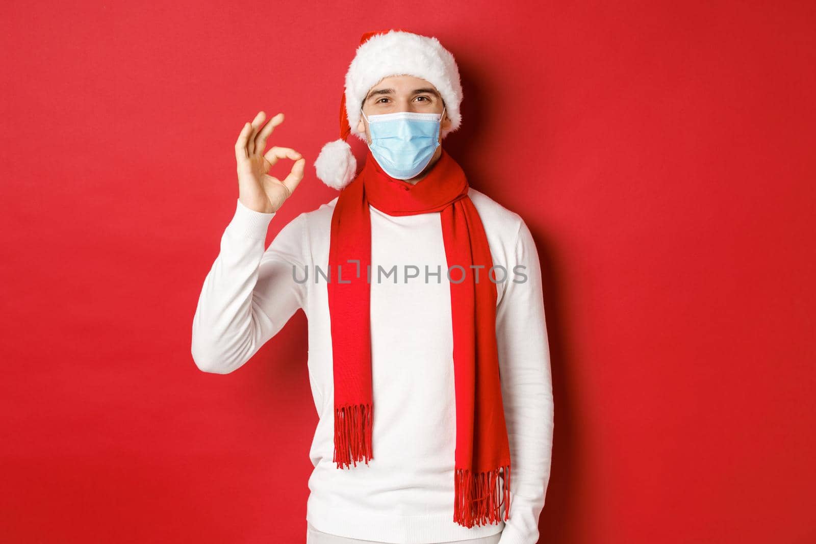 Concept of covid-19, christmas and holidays during pandemic. Portrait of happy and satisfied man in medical mask and santa hat, showing okay sign in approval, recommending something.