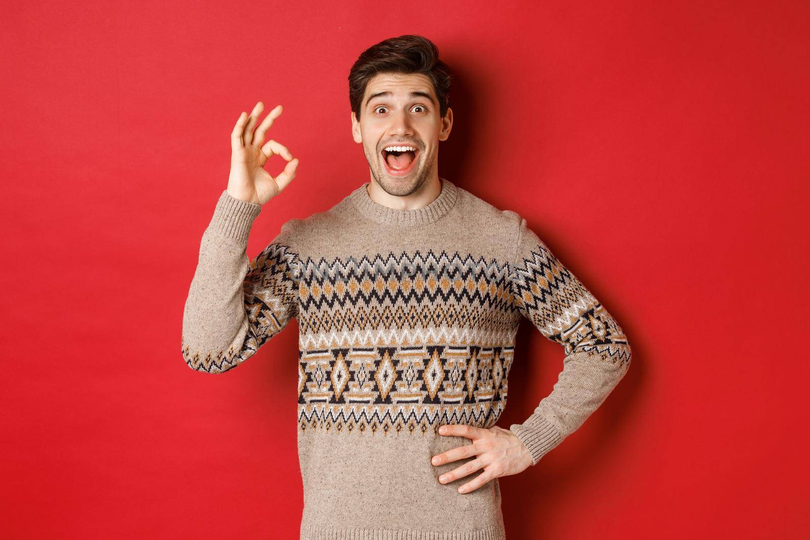 Concept of christmas celebration, winter holidays and lifestyle. Amazed and happy man in xmas sweater, showing okay sign and looking at something fantastic, red background.