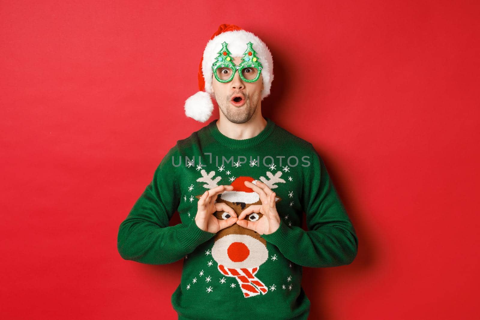 Portrait of carefree handsome man in santa hat and party glasses, making fun of his christmas sweater, looking happy over red background.