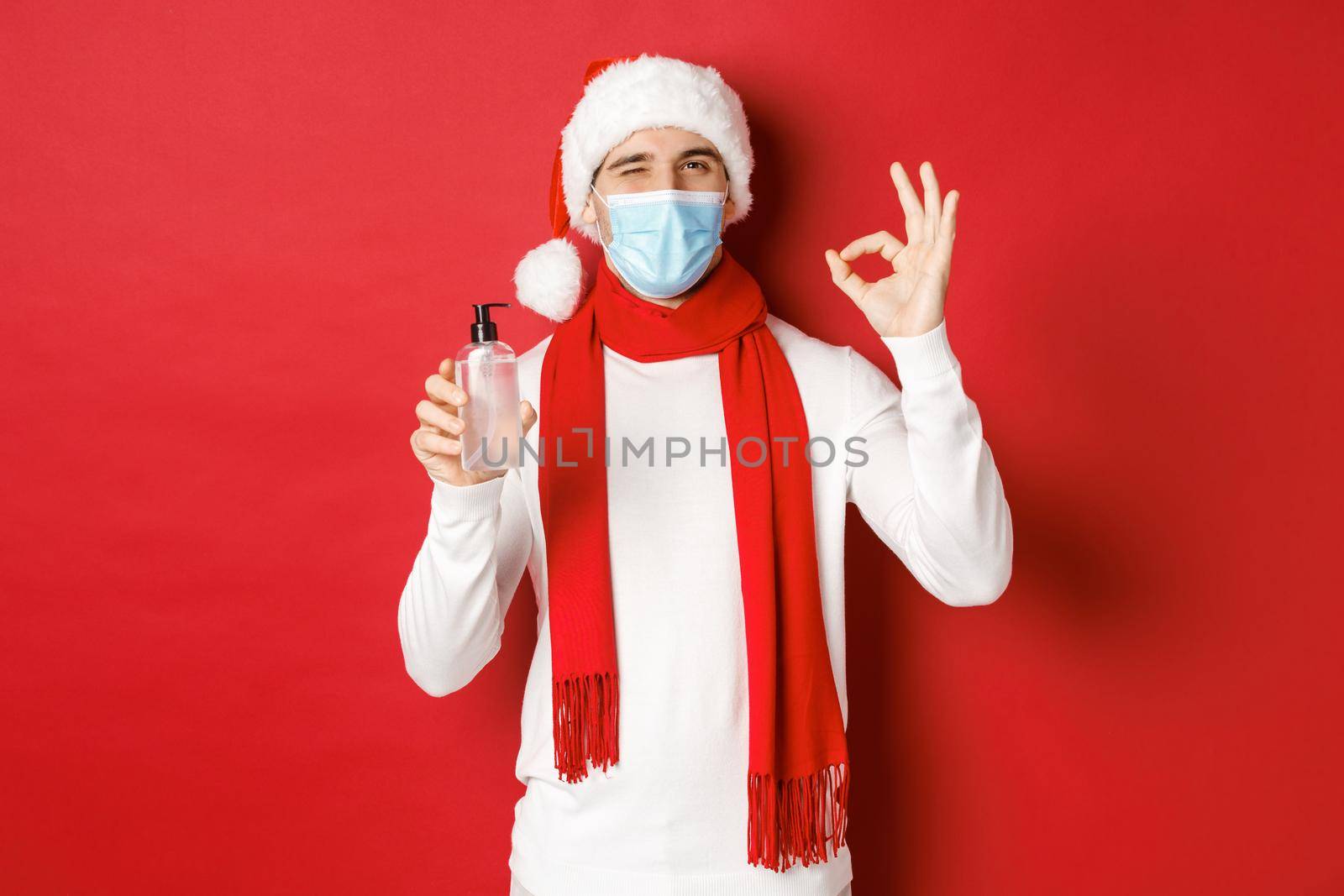 Concept of covid-19, christmas and holidays during pandemic. Cheerful man celebrating new year in medical mask and santa hat, recommending hand sanitizer, showing okay sign and winking.