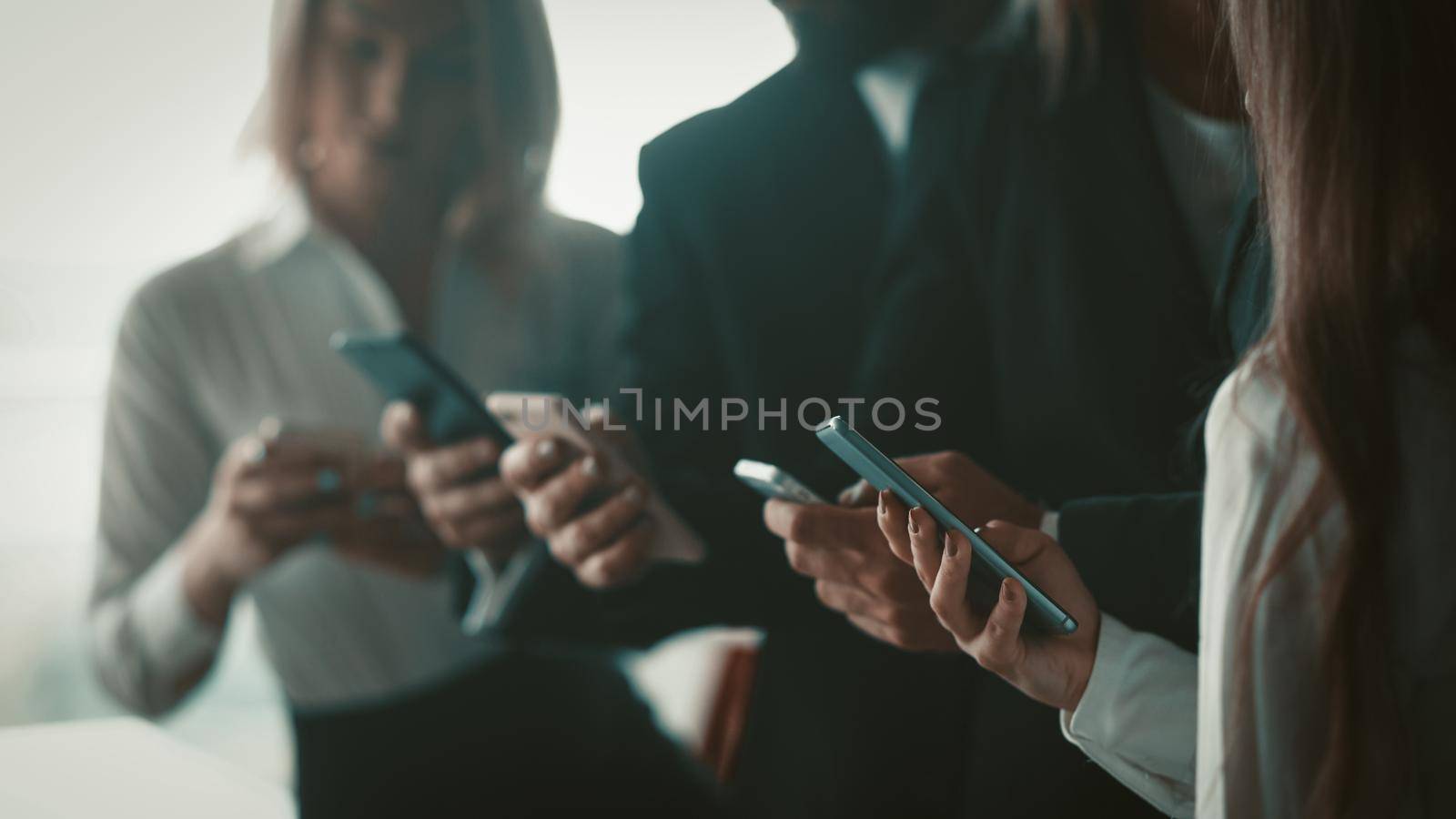 Business people using mobile phones together standing in office. Close up shot of human hands holding smartphones. Blurred image. Selective focus on female hand in foreground.