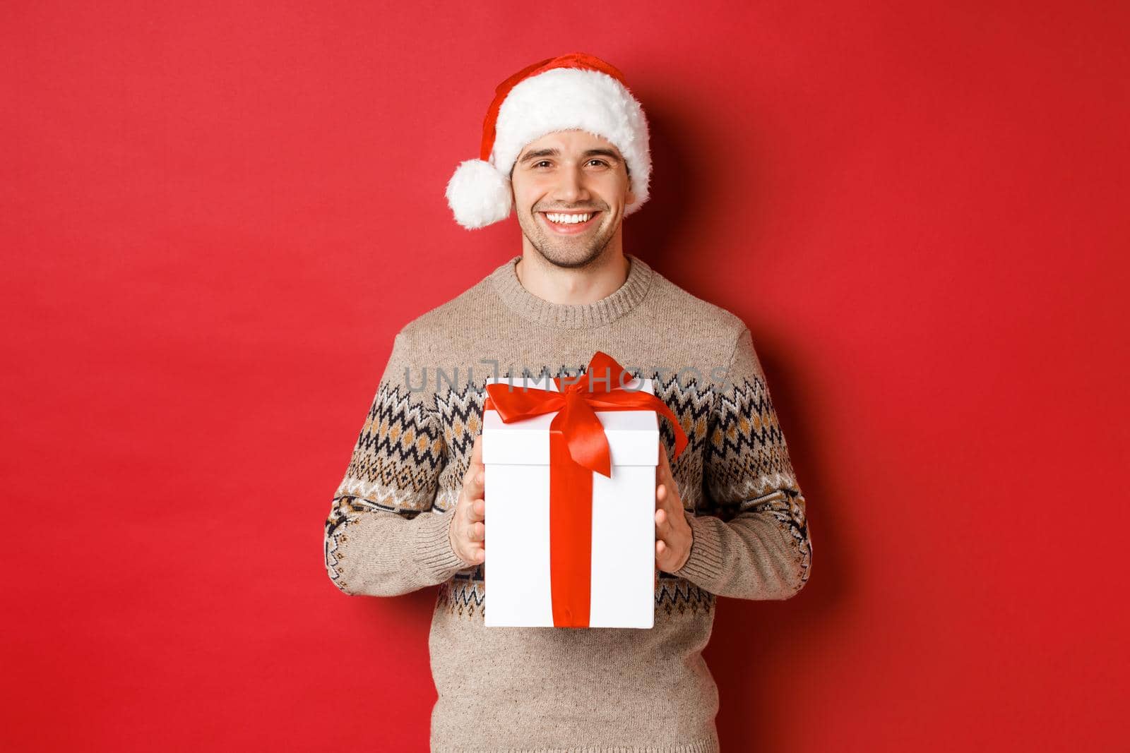 Image of handsome smiling man in santa hat and winter sweater, holding a present, giving christmas gift and wishing happy holidays, standing over red background.