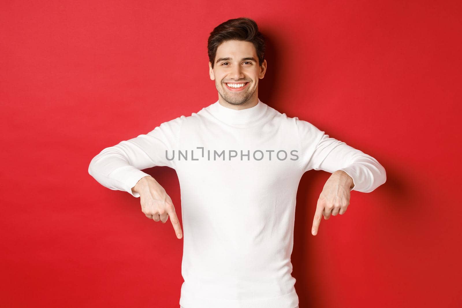 Image of smiling handsome man in white sweater inviting visit page, pointing fingers down and showing christmas banner, standing against red background.