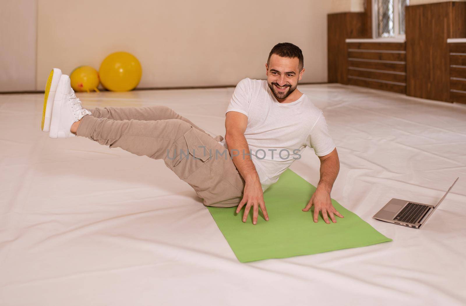 High top angle. Young handsome man doing crunches abs core looking at camera while playing online work out videos on using laptop. Young man warming up doing special exercises for abs muscles.