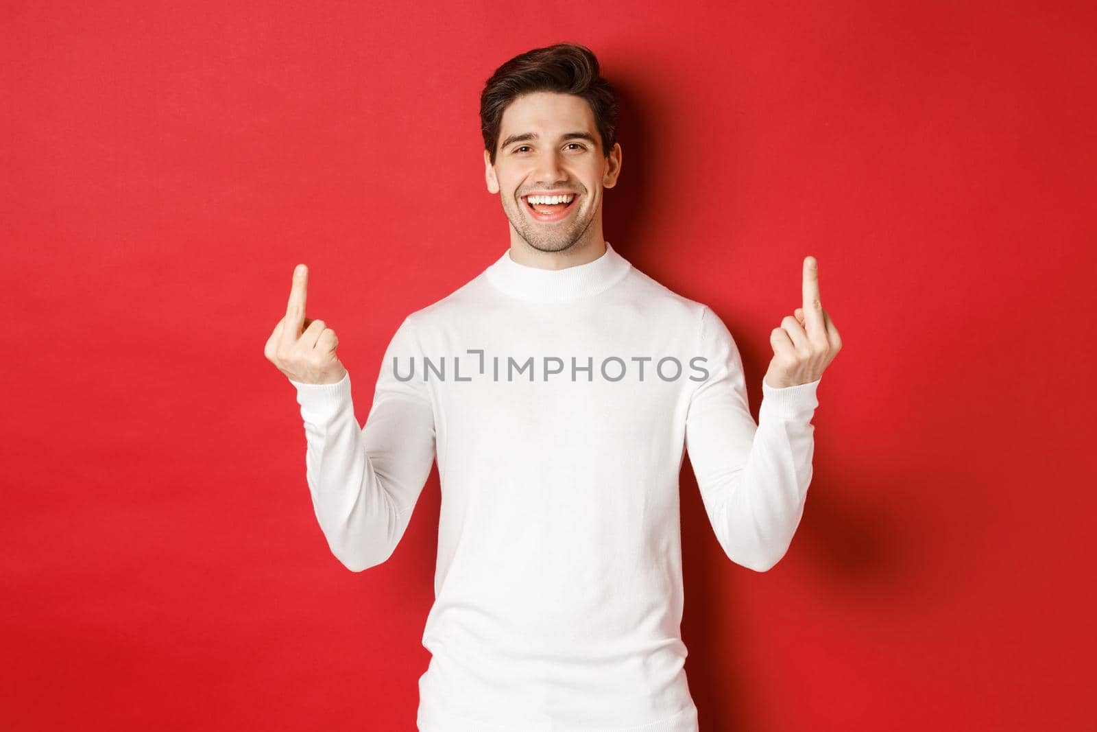 Image of rude and unbothered man laughing while showing middle-fingers, telling to fuck-off, standing over red background.