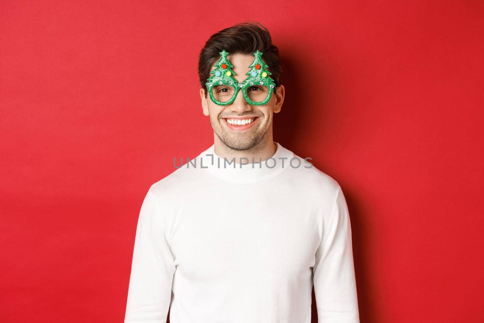 Concept of christmas, winter holidays and celebration. Close-up of handsome man in white sweater and party glasses, enjoying new year, standing over red background by Benzoix