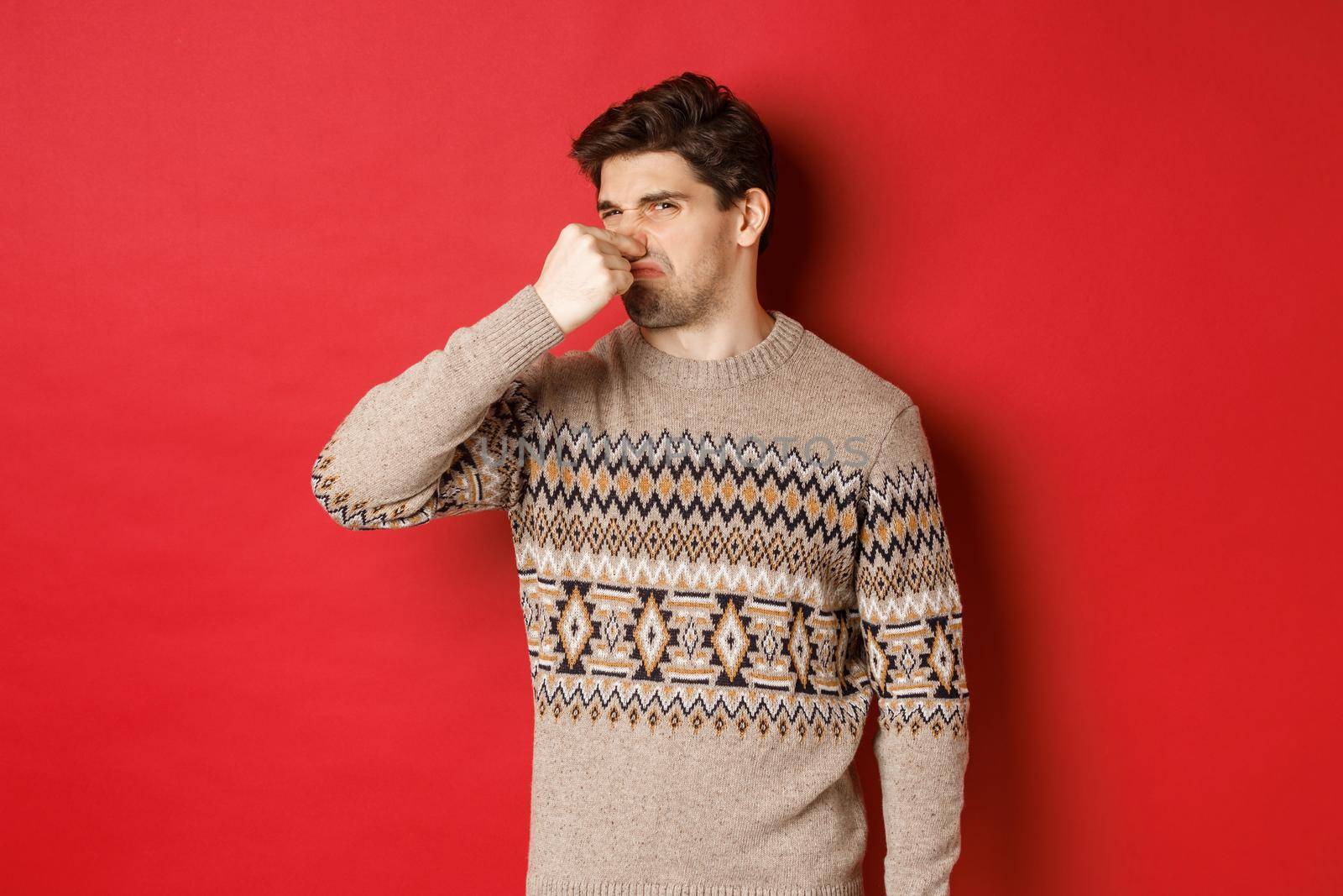 Image of disgusted young man in winter sweater, shut nose and grimacing from bad smell, standing over red background by Benzoix