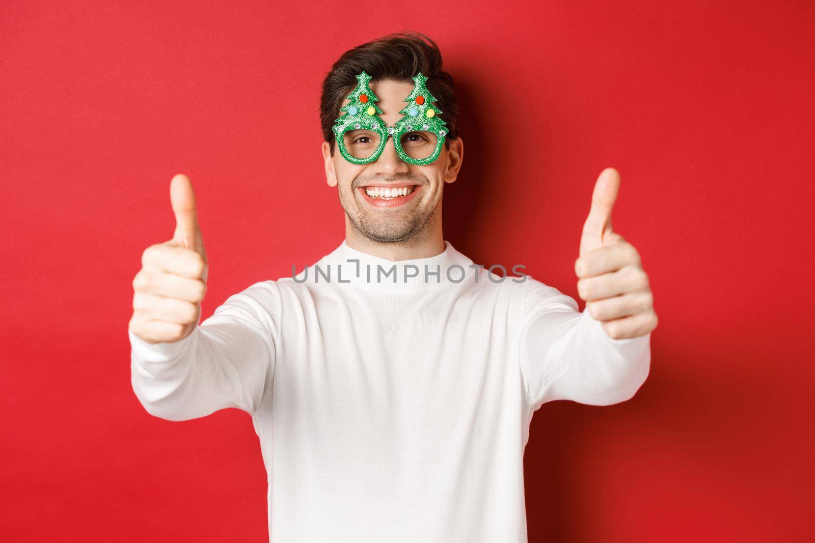 Close-up of attractive happy man in party glasses and white sweater, showing thumbs-up in approval and smiling, standing over red background by Benzoix