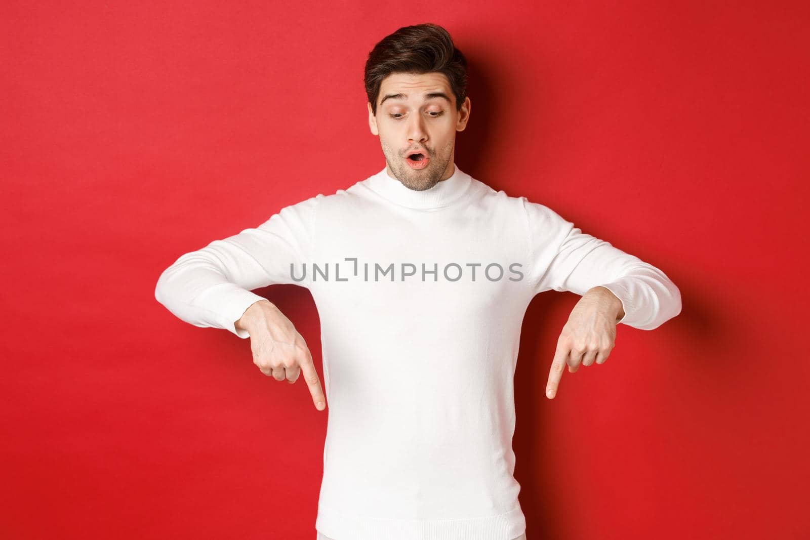 Portrait of surprised good-looking guy in white sweater, looking and pointing fingers down at logo, standing against red background.