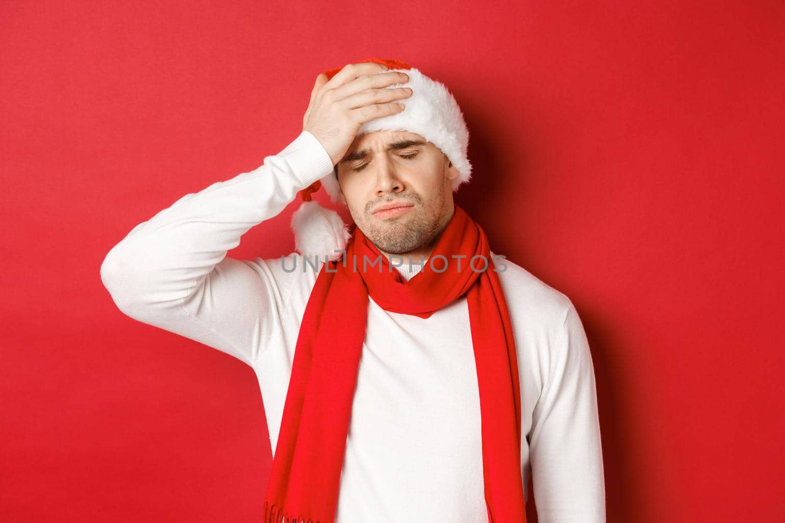 Concept of christmas, winter holidays and celebration. Portrait of sad and frustrated man in santa hat, feeling bad, standing over red background by Benzoix