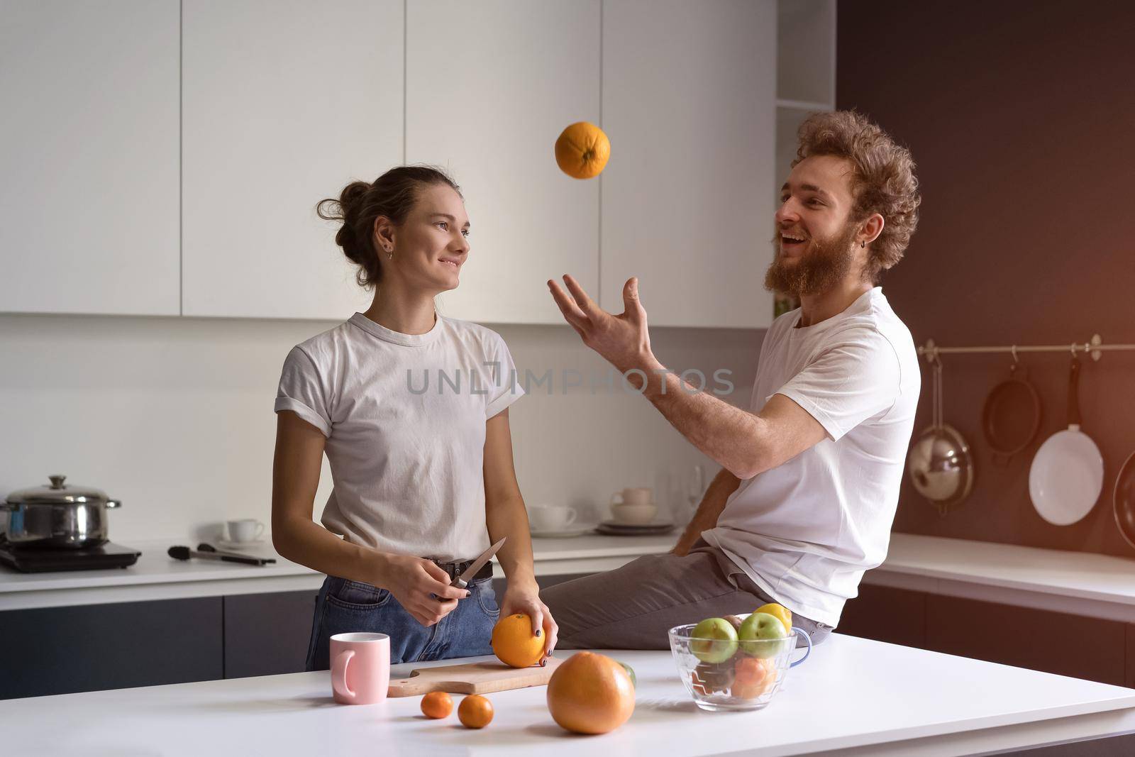 Playing with orange young man happy of their new home young couple cooking in new kitchen. Beautiful young couple talking enjoying their new house. New home concept by LipikStockMedia