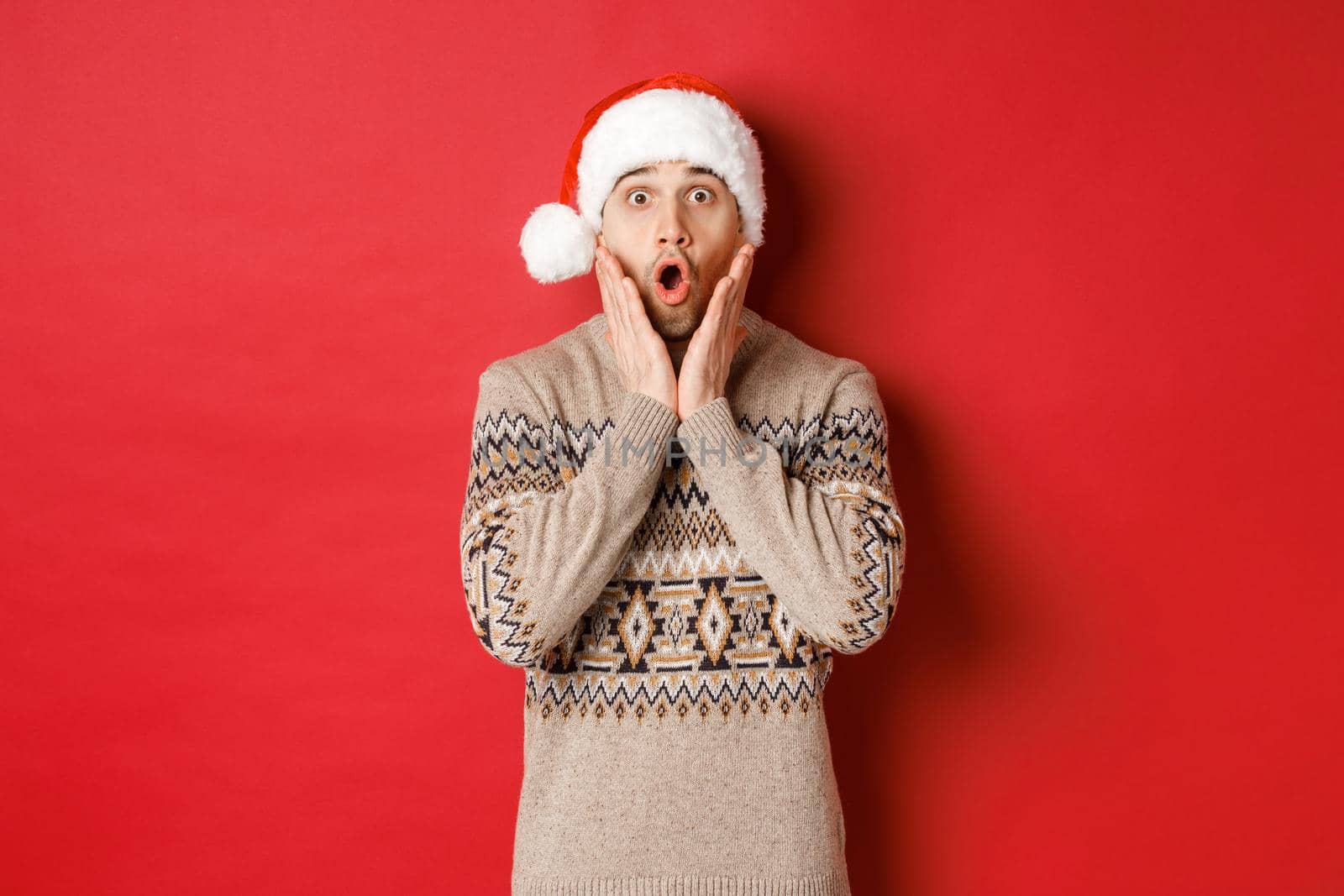 Image of amazed and speechless man in santa hat, drop jaw and staring at awesome christmas offer, standing over red background by Benzoix