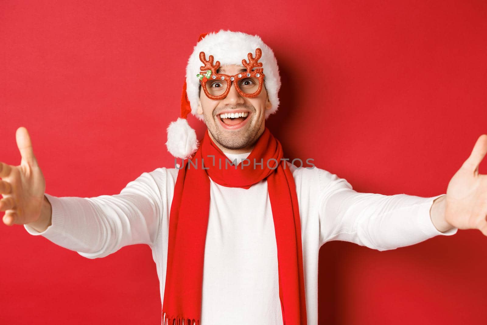Concept of christmas, winter holidays and celebration. Close-up of happy smiling man in party glasses and santa hat, spread hands sideways for hug, greeting guests, red background.