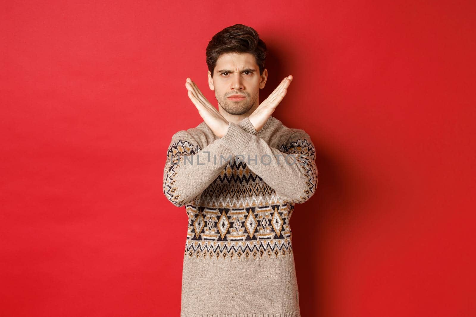 Image of angry and serious handsome man in christmas sweater, telling no or stop, showing cross gesture to restrict you from something, prohibit action, standing over red background.