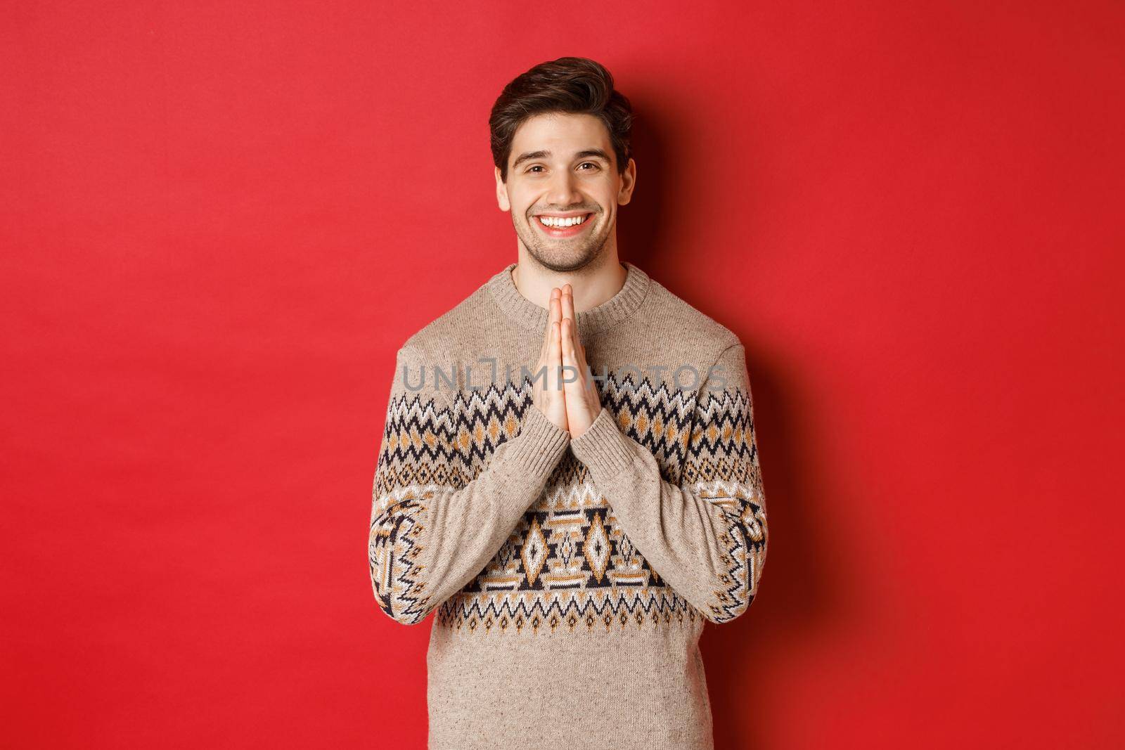 Image of happy and cute man in christmas sweater, asking for favour, holding hands together, smiling and saying thank you, feeling grateful, standing over red background.