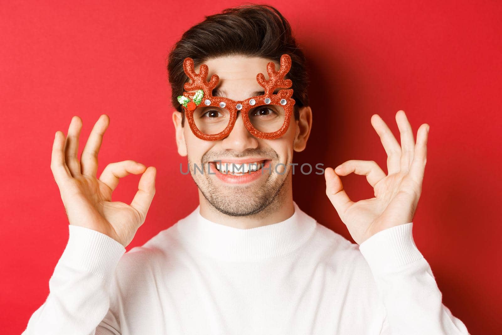 Concept of winter holidays, christmas and celebration. Close-up of attractive man in party glasses, showing okay signs and smiling, praise something good, red background by Benzoix