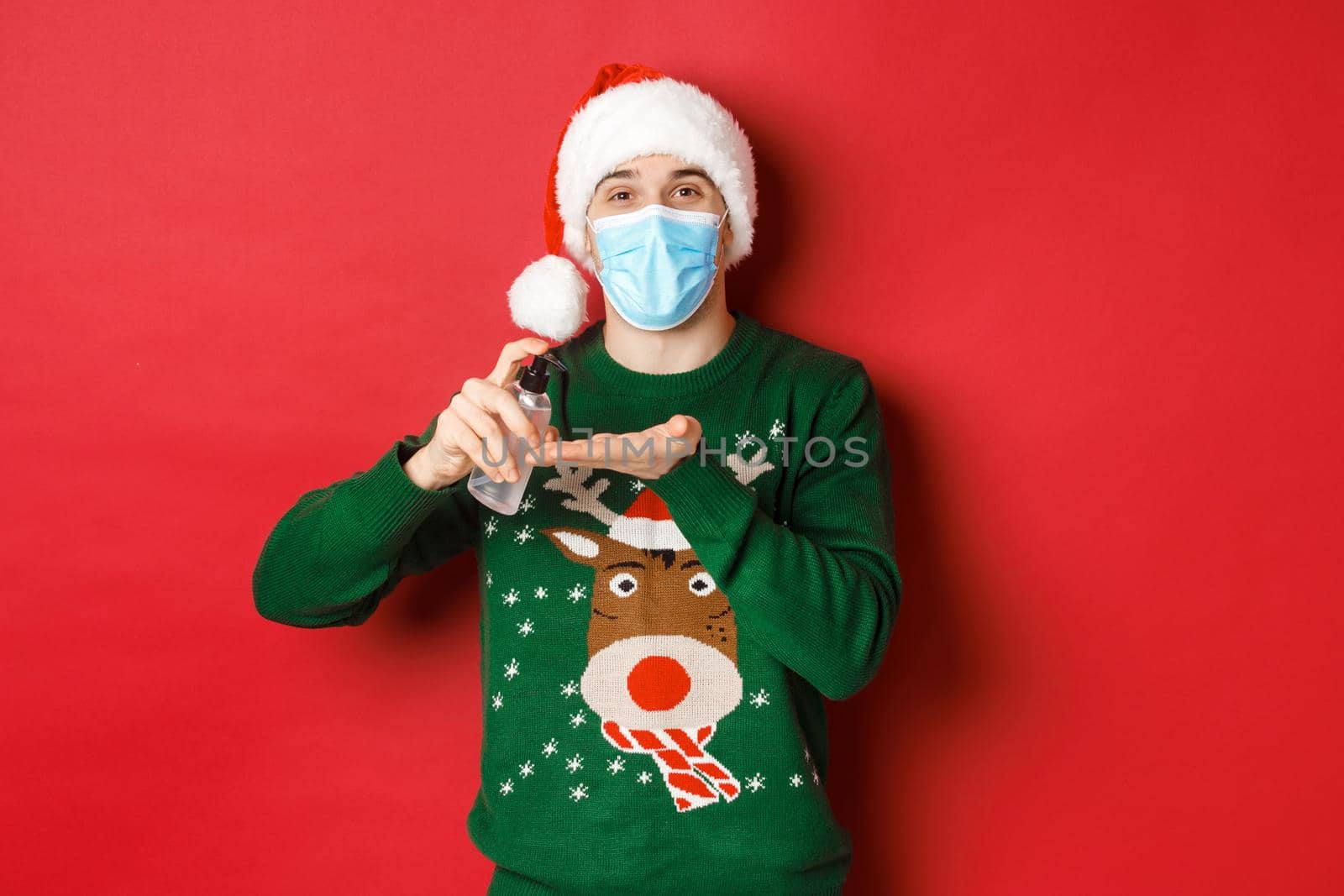 Concept of new year, coronavirus and social distancing. Attractive young man in santa hat, medical mask and christmas sweater, using hand sanitizer to clean hands, standing over red background.
