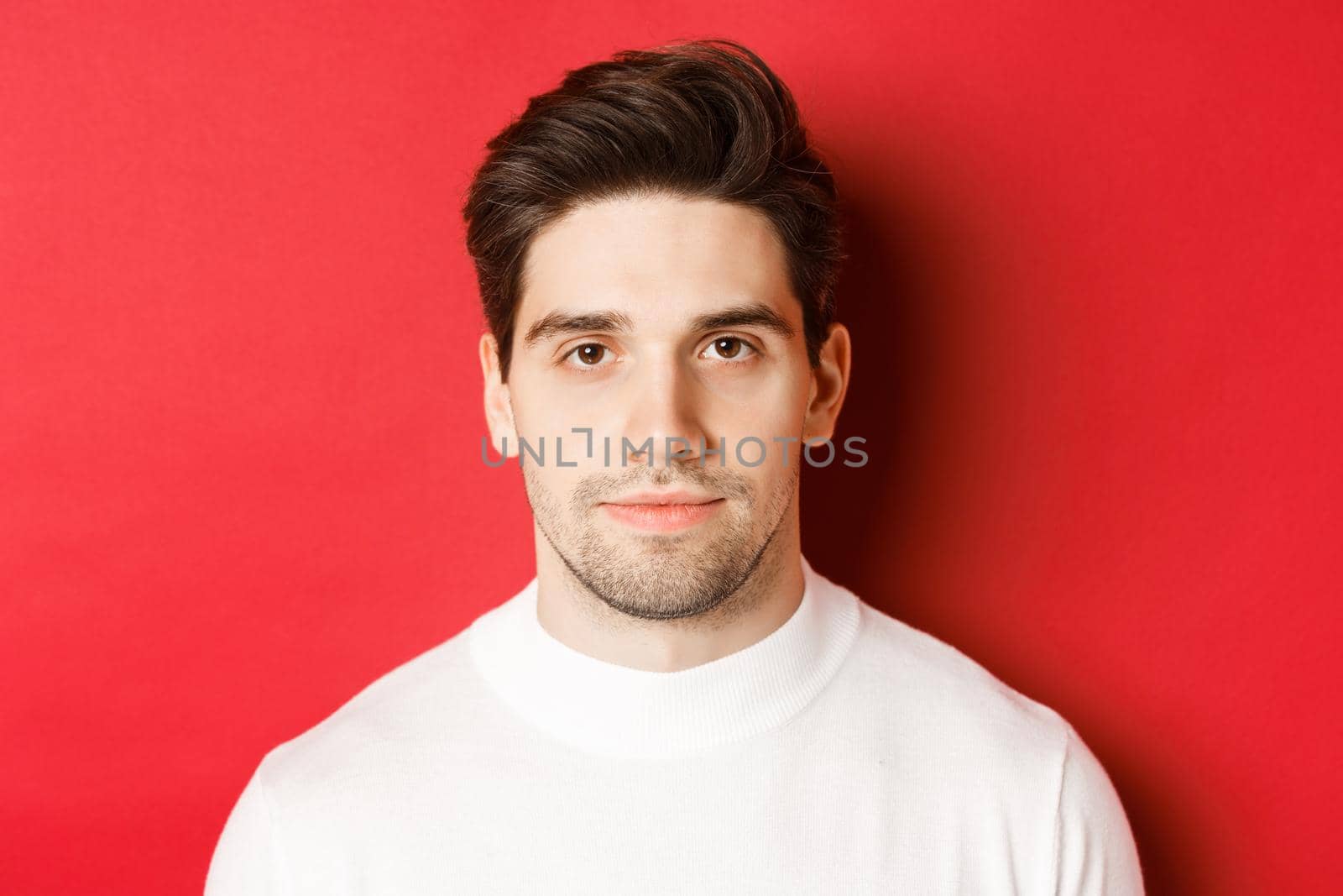 Close-up of handsome brunette guy in white sweater, smiling at camera, standing over red background.