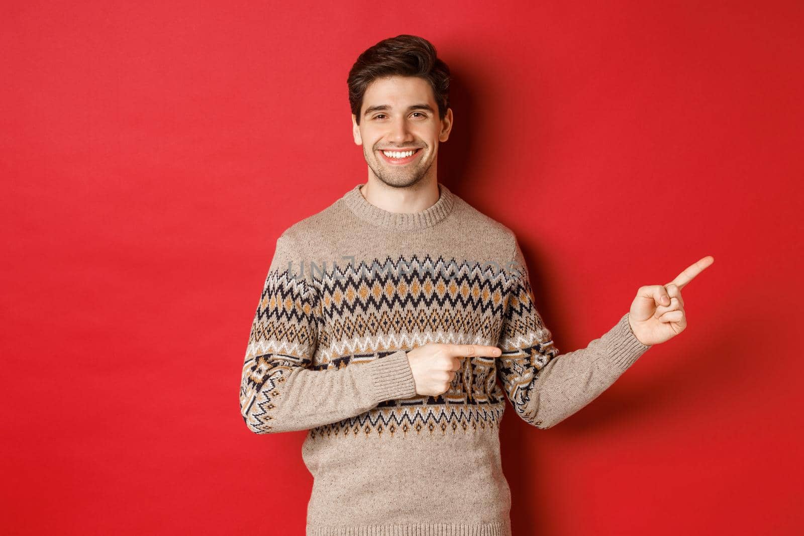 Image of attractive smiling man, wearing christmas sweater, pointing fingers right and showing new year advertisement, standing over red background.