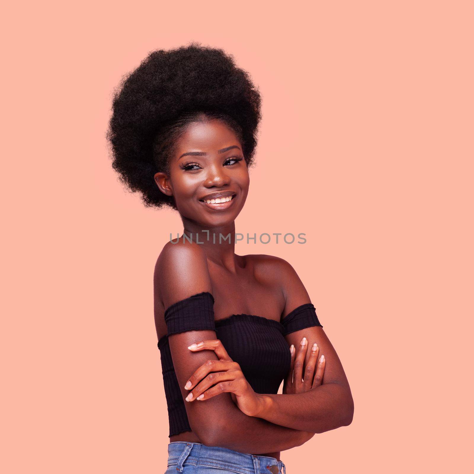 Square portrait of happy charming African American girl with amazing afro hairstyle smiling with arms folded. Dark-skinned lady wearing black bare shoulders top and denim jeans on peach background.