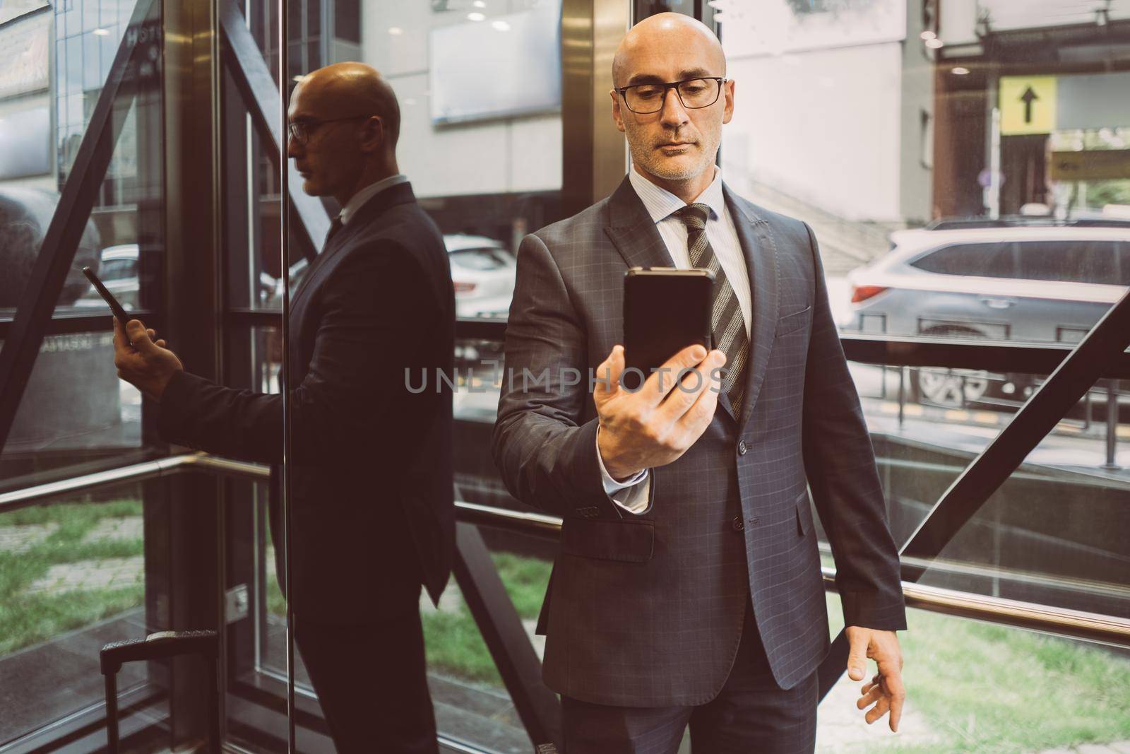 Thoughtful businessman in suit reading information in smartphone hold in it in front of him standing next to a window with a busy street view. Focused business man face with smartphone.