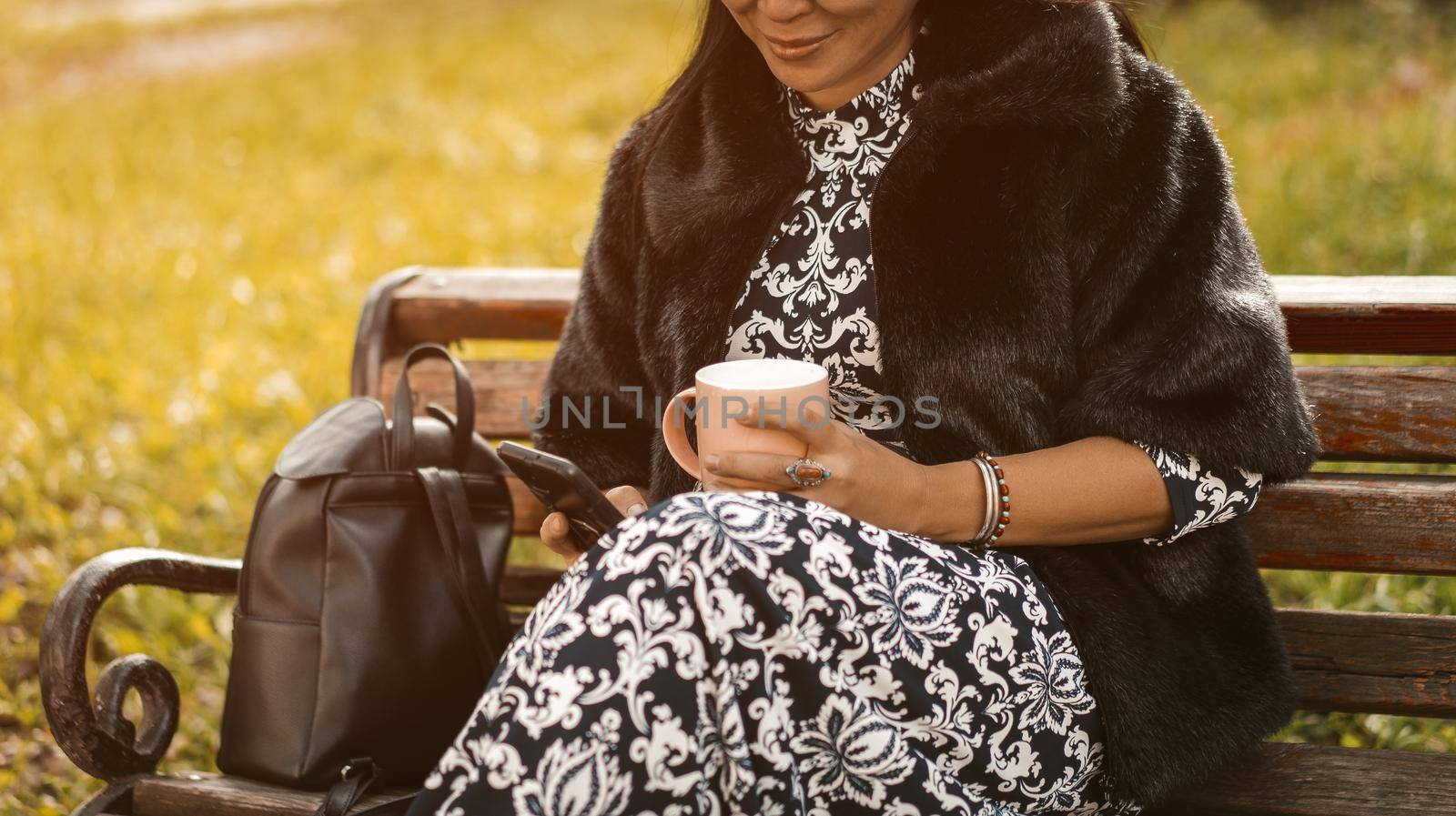 Asian woman texting on the smartphone while drinking coffee wearing fur coat and dress sits at park on old rusty bench. Resting woman in sunny day outdoors. Close up.