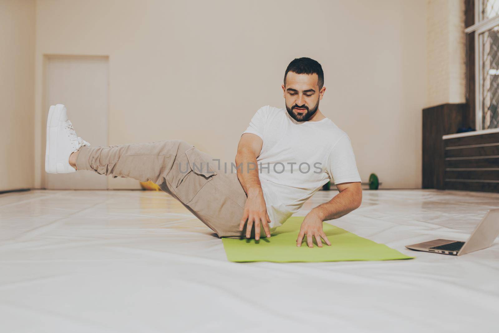 Low angle. Young handsome man doing crunches and looking online work out videos on using laptop. Self motivated young man warming up doing special exercises for abs muscles by LipikStockMedia