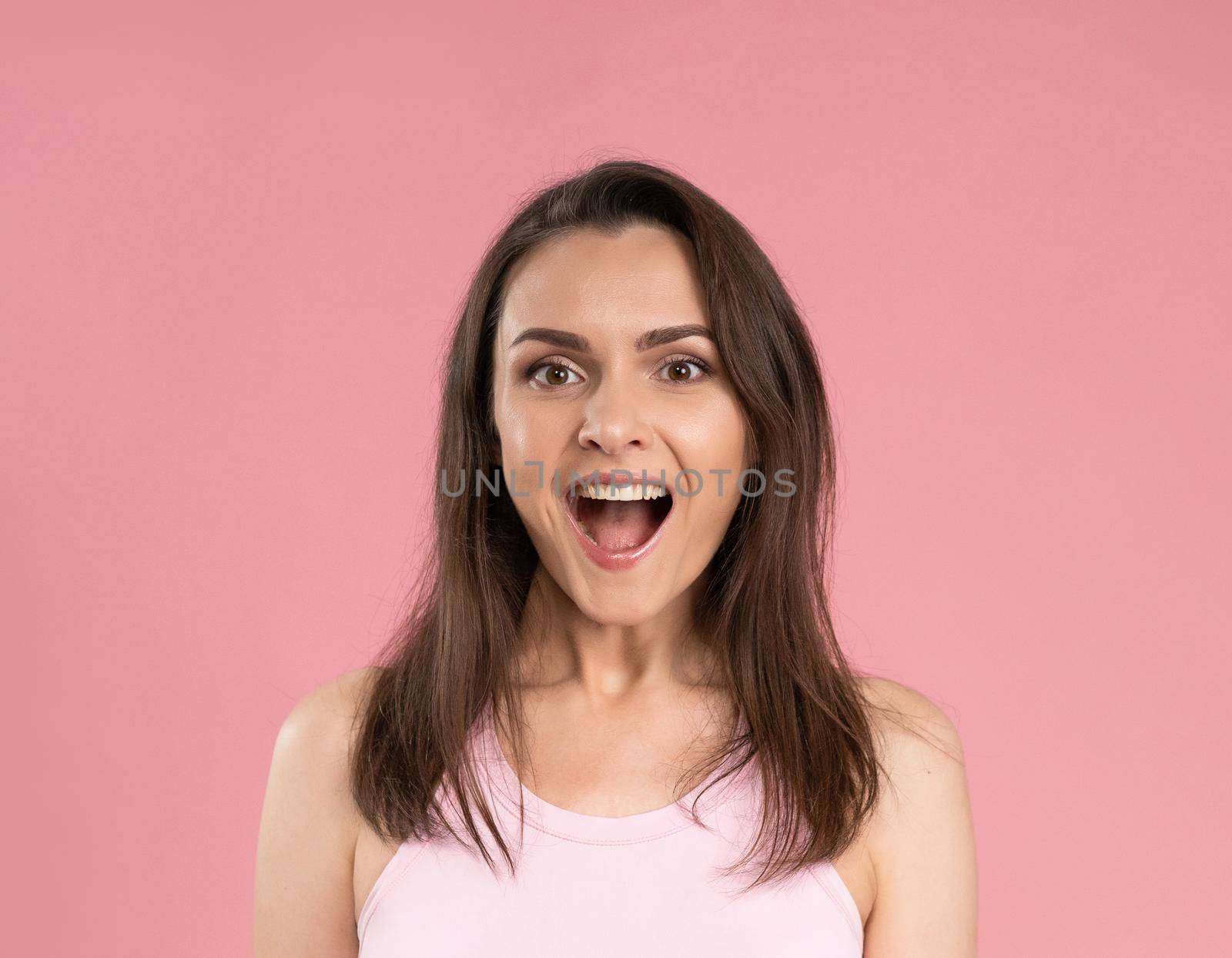 Beautiful young female emotional smiling and surprised looking at camera. Young woman with dark hair in pink t-shirt looking at camera with mouth open. Facial expressions, emotions, feelings.