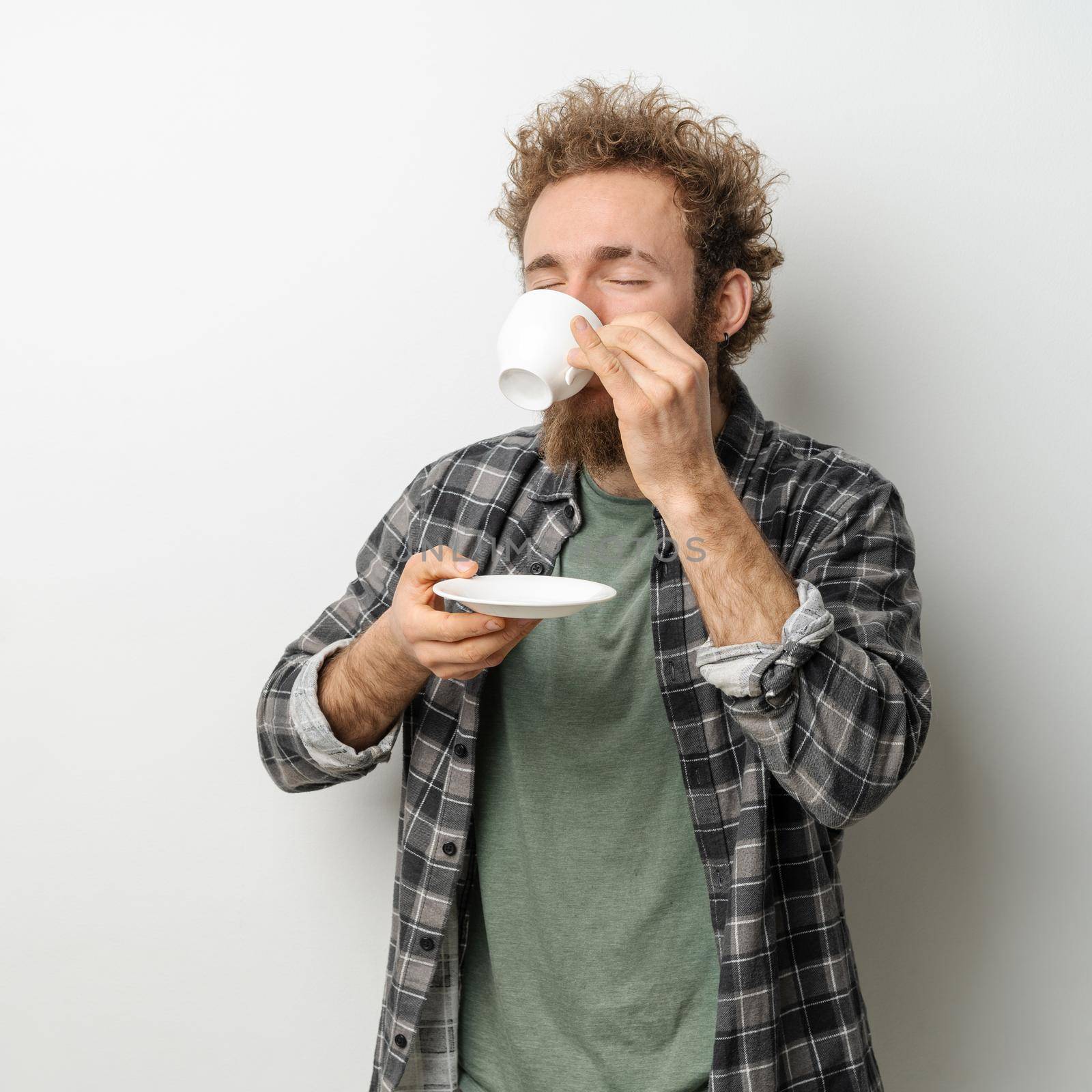 Good looking man with curly hair and beard drinking coffee holding cup, wearing plaid long sleeve shirt isolated on white background.