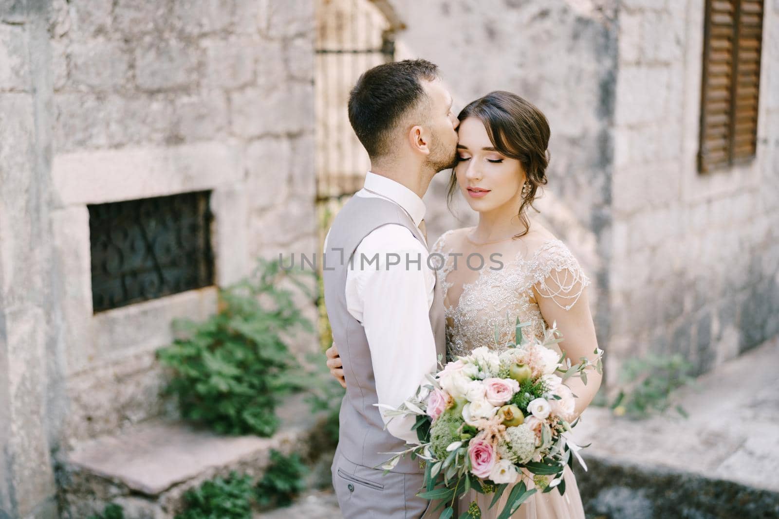 Groom kisses bride forehead near the stone building. Portrait. High quality photo