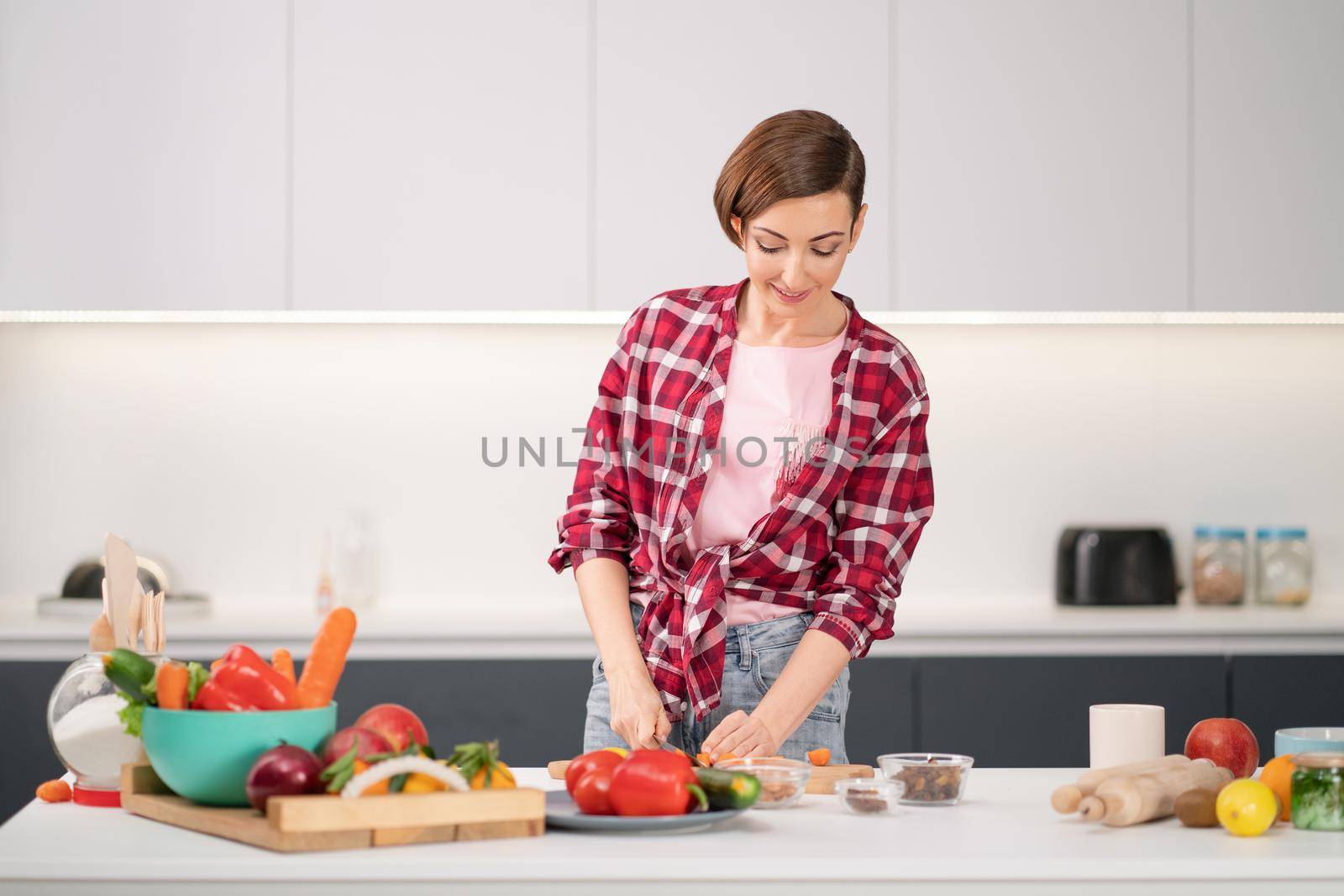 Pretty housewife cutting fresh carrot cooking dinner for her family wearing a plaid shirt. Cooking with passion young woman with short hair standing at modern kitchen. Healthy food leaving - concept by LipikStockMedia