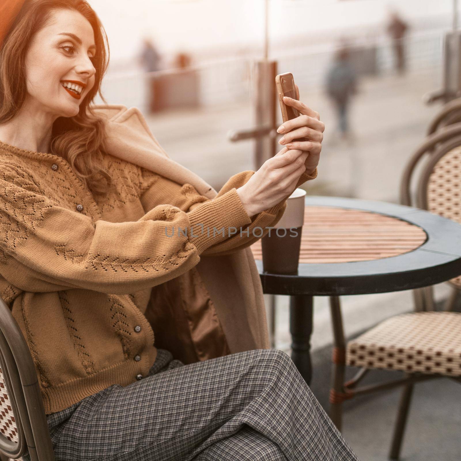 Beautiful young woman video calling using her smartphone while having cup of coffee sitting in a spring outdoor cafe. French woman in red beret with background of urban city. Square cropped by LipikStockMedia