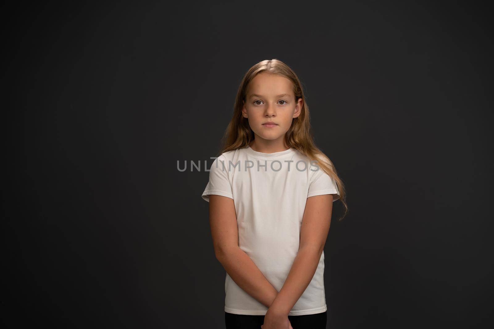 Little girl standing looking questioningly or frustrated at the camera wearing white t-shirt isolated on dark grey or black background.