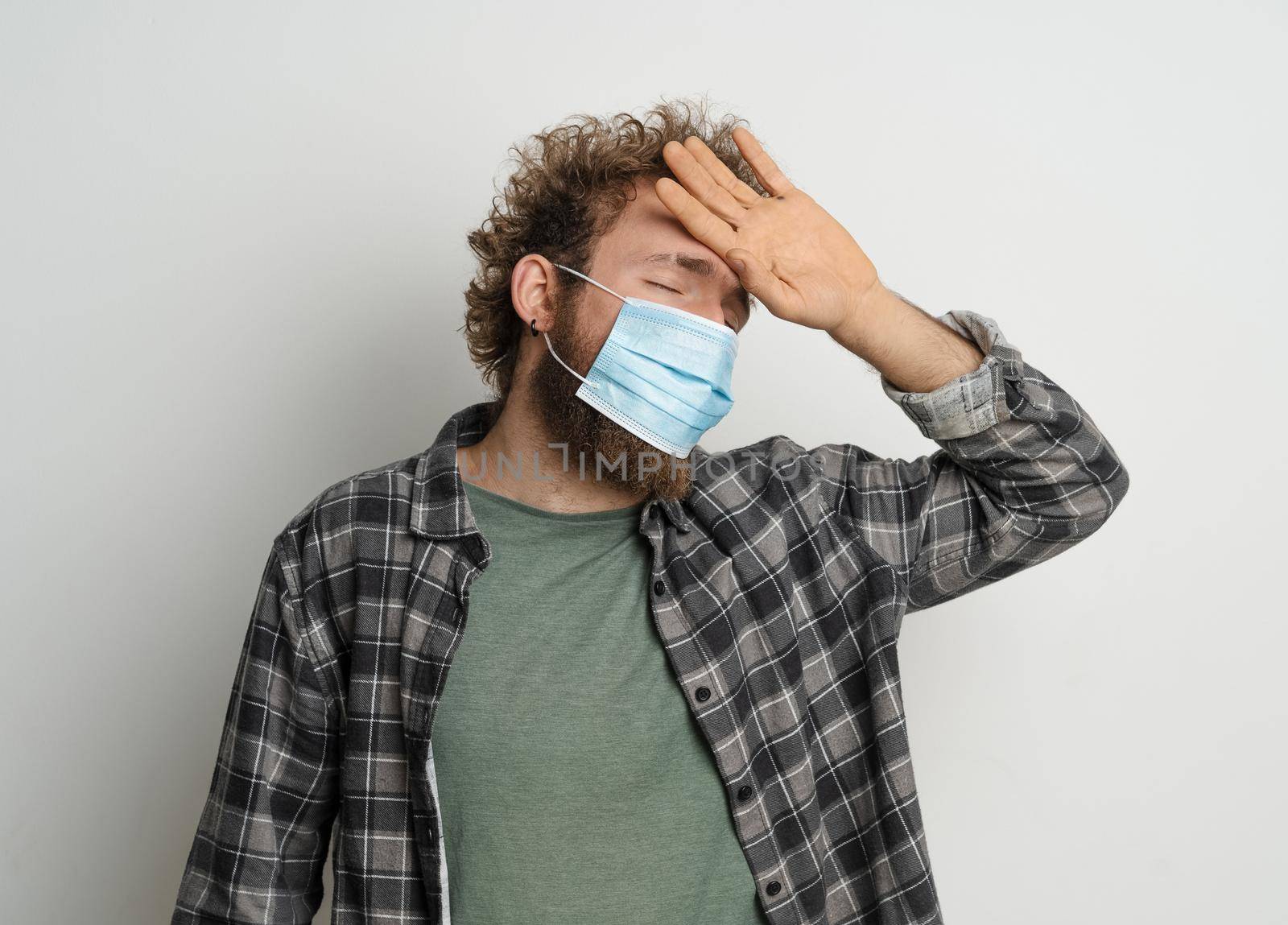Feeling high temperature young man wearing protective sterile medical mask on his face to protect coronavirus curly hair in plaid shirt and olive t-shirt under. Medicine concept. White background.
