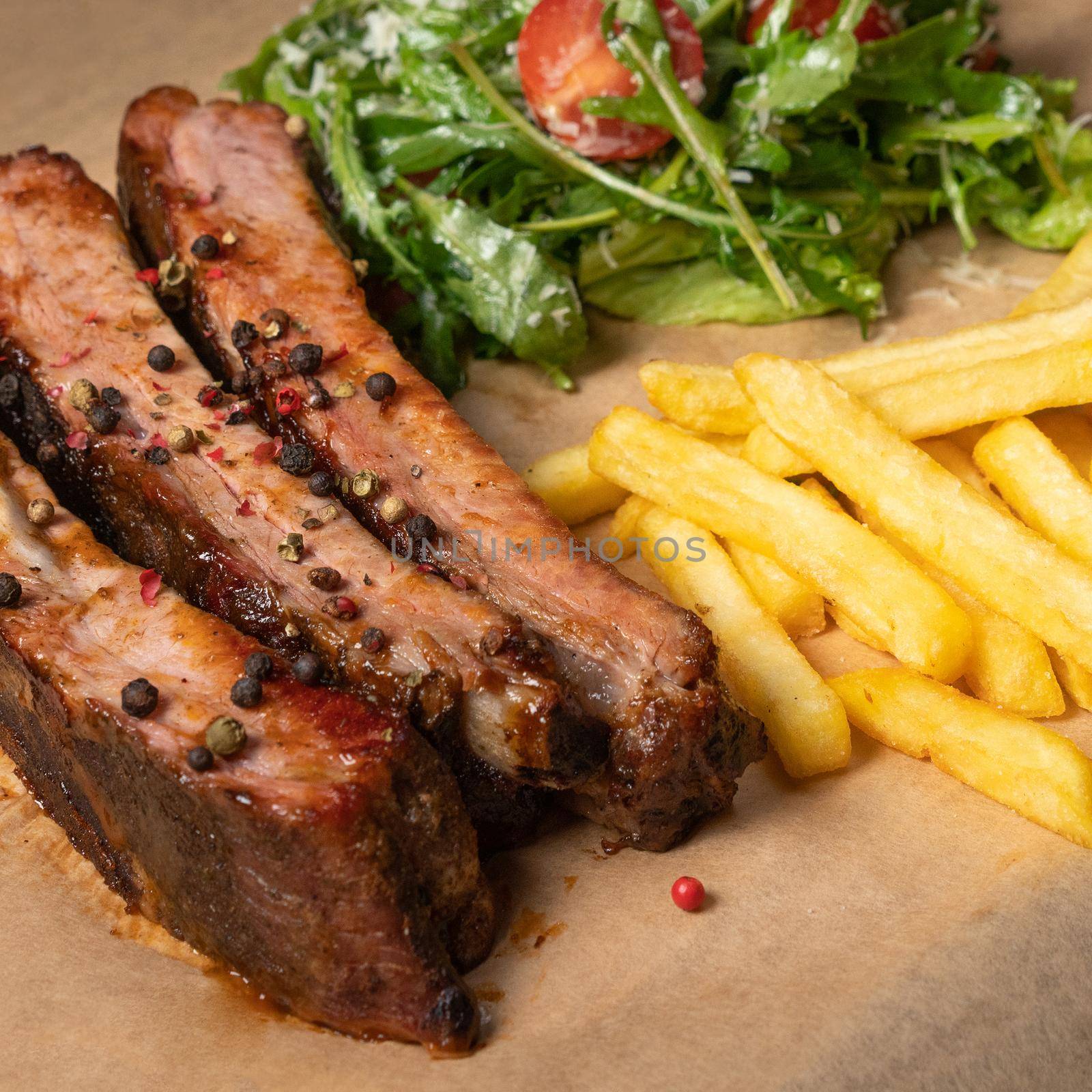 BBQ spare ribs served with French fries and arugula salad with cherry tomatoes and parmesan cheese served on board covered with food grade paper. Restaurant concept. Square cropped image by LipikStockMedia