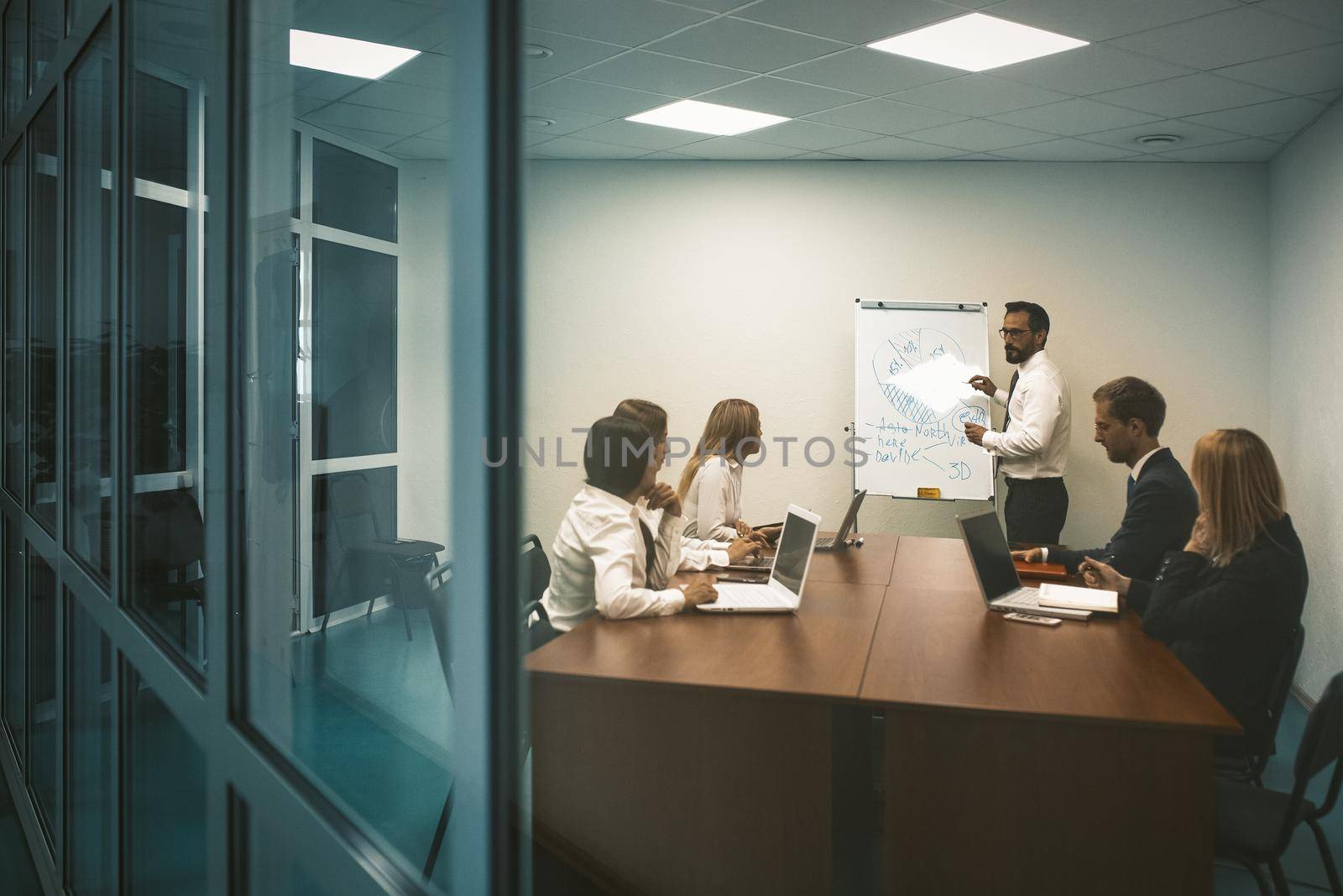 Business presentation concept. Speaker man stands near whiteboard having discussion with business people colleagues who are sitting at office table in office by LipikStockMedia