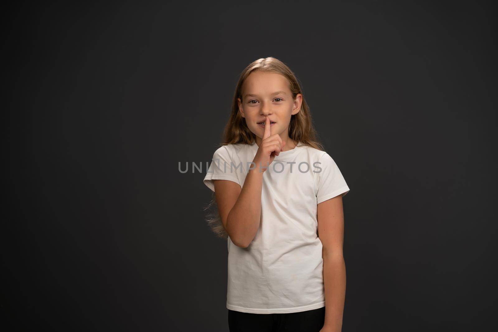 Little girl holds her finger on her lips telling everyone to keep silence. Child wearing white t shirt smiling at the camera isolated on dark grey or black background.