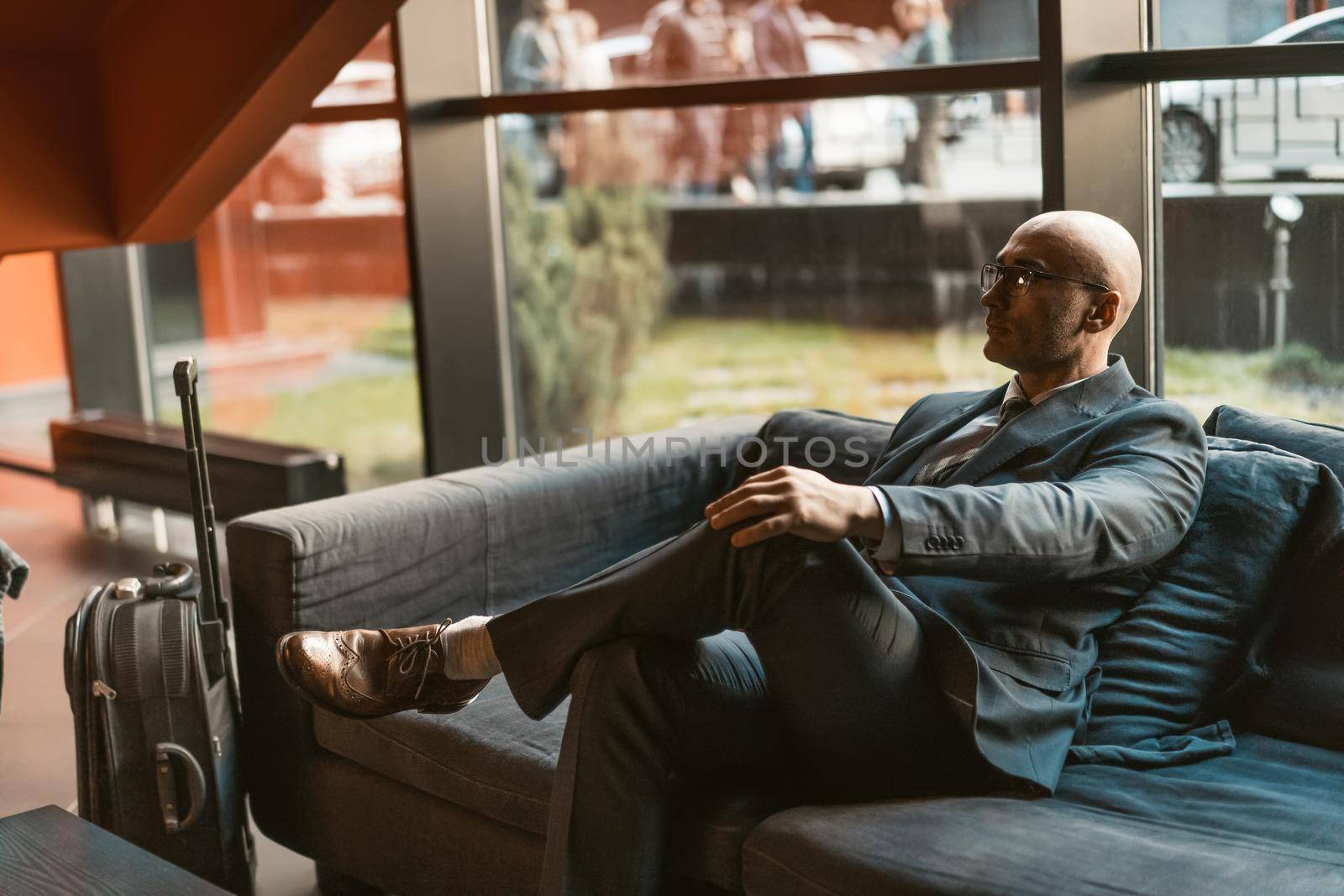 Waiting in hotel lobby respectable businessman looking away while waiting for check in or check out attending business forum. Handsome wearing business suit sitting in dark sofa in reception area.