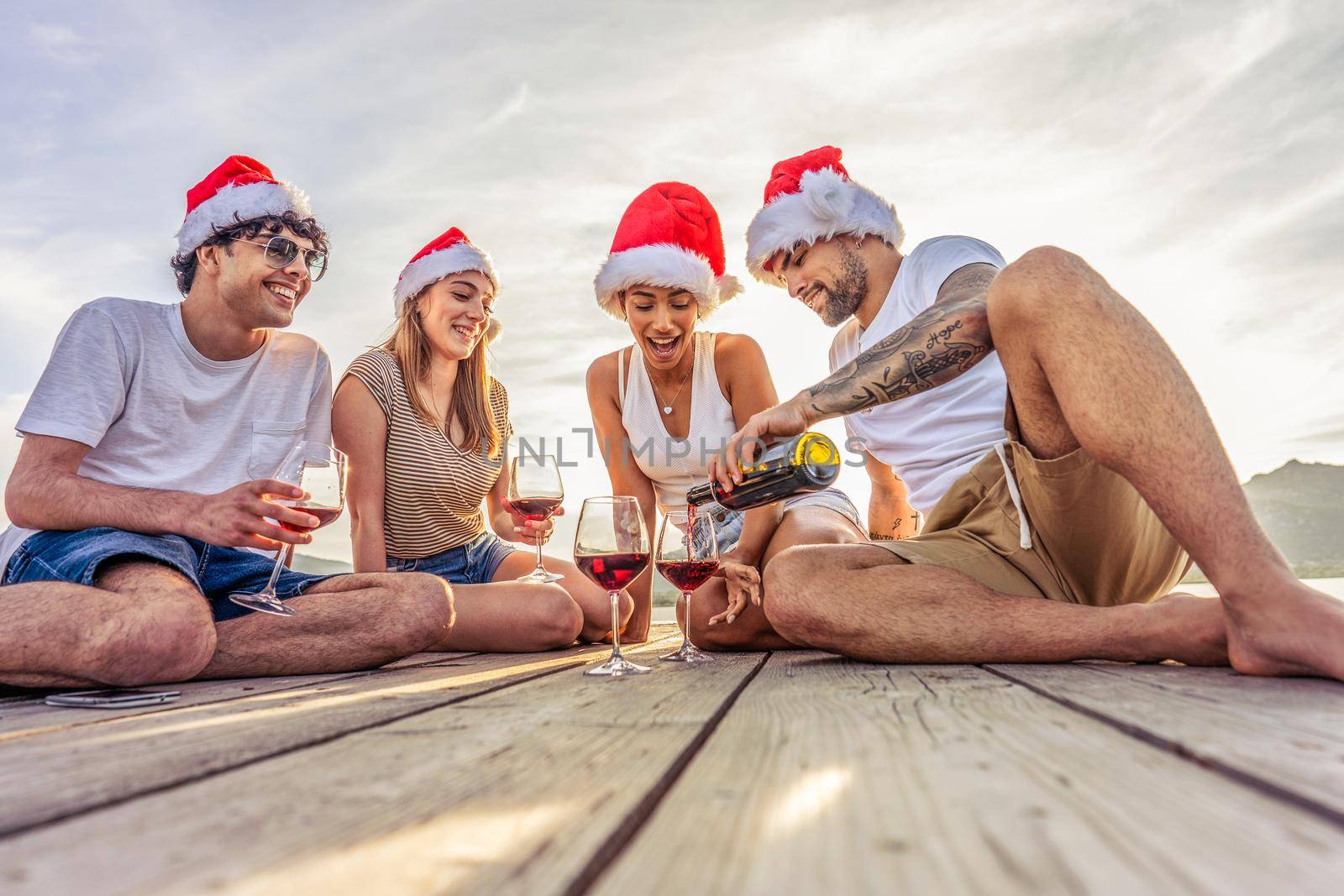 Happy people enjoying outdoor life in exotic vacation resort drinking alcohol. Group of young friends having fun together at sunset wearing red Santa Claus hat sitting on a pier in sea vacation resort by robbyfontanesi