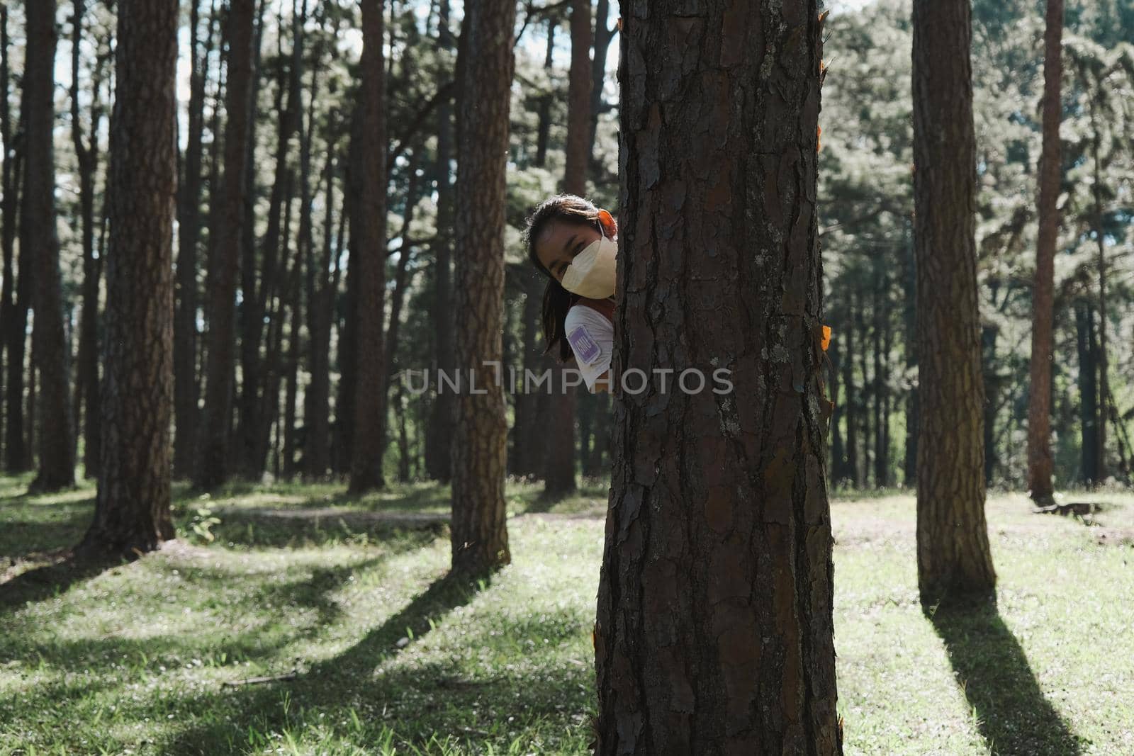 Smiling woman in mask peeking out from behind tree in a park. Happy woman hiding behind a tree in a summer day. Love nature concept.