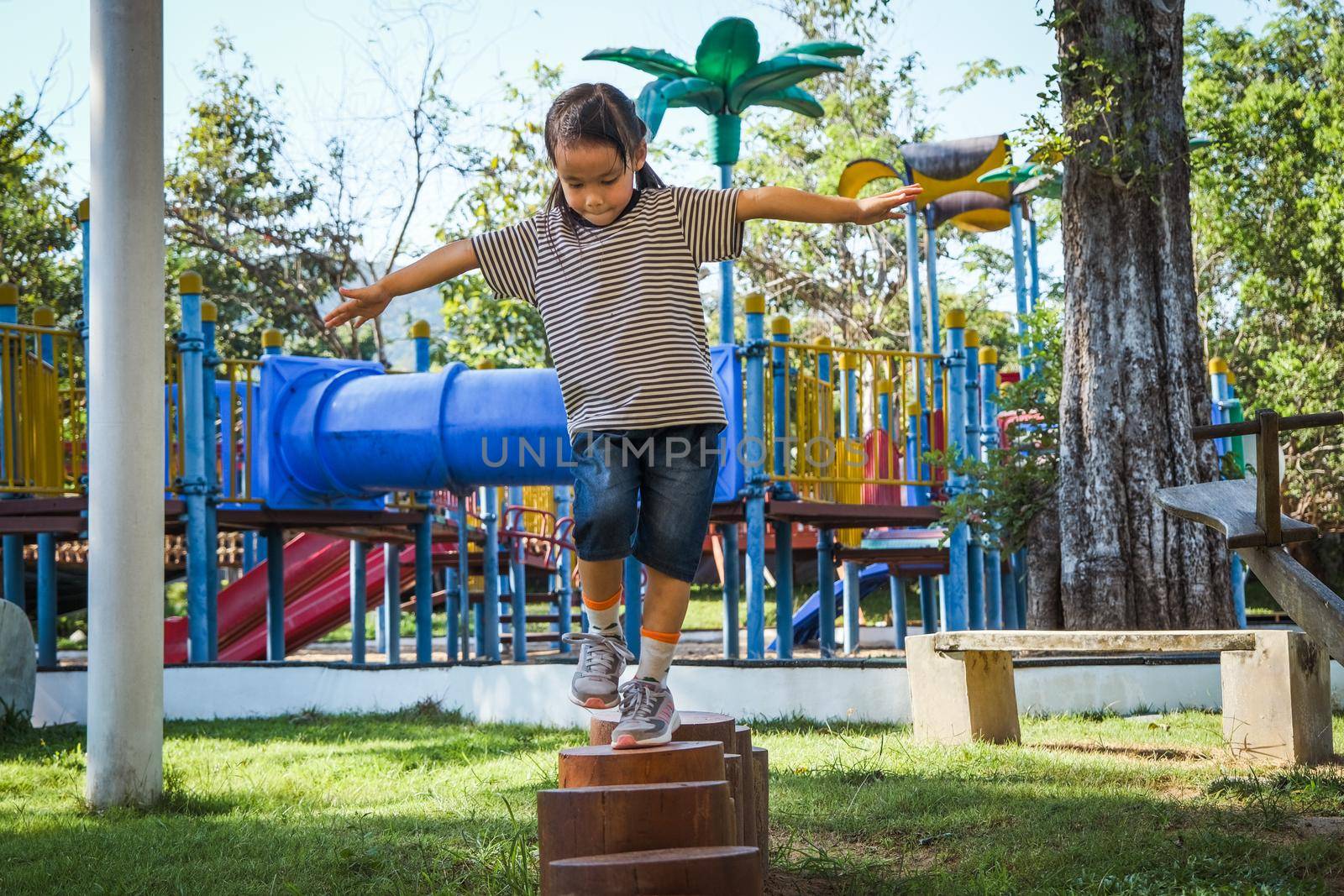 Active little girl walking on balance beam in the outdoor playground in the park. Happy child girl having fun in children playground. Play is learning in childhood. by TEERASAK