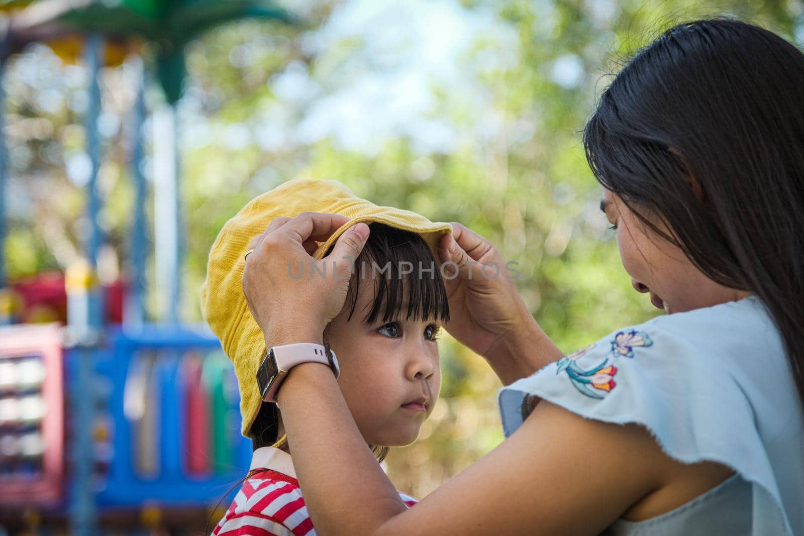 Young mother puts a hat on her daughter in the summer park. Family spends time together on vacation. by TEERASAK