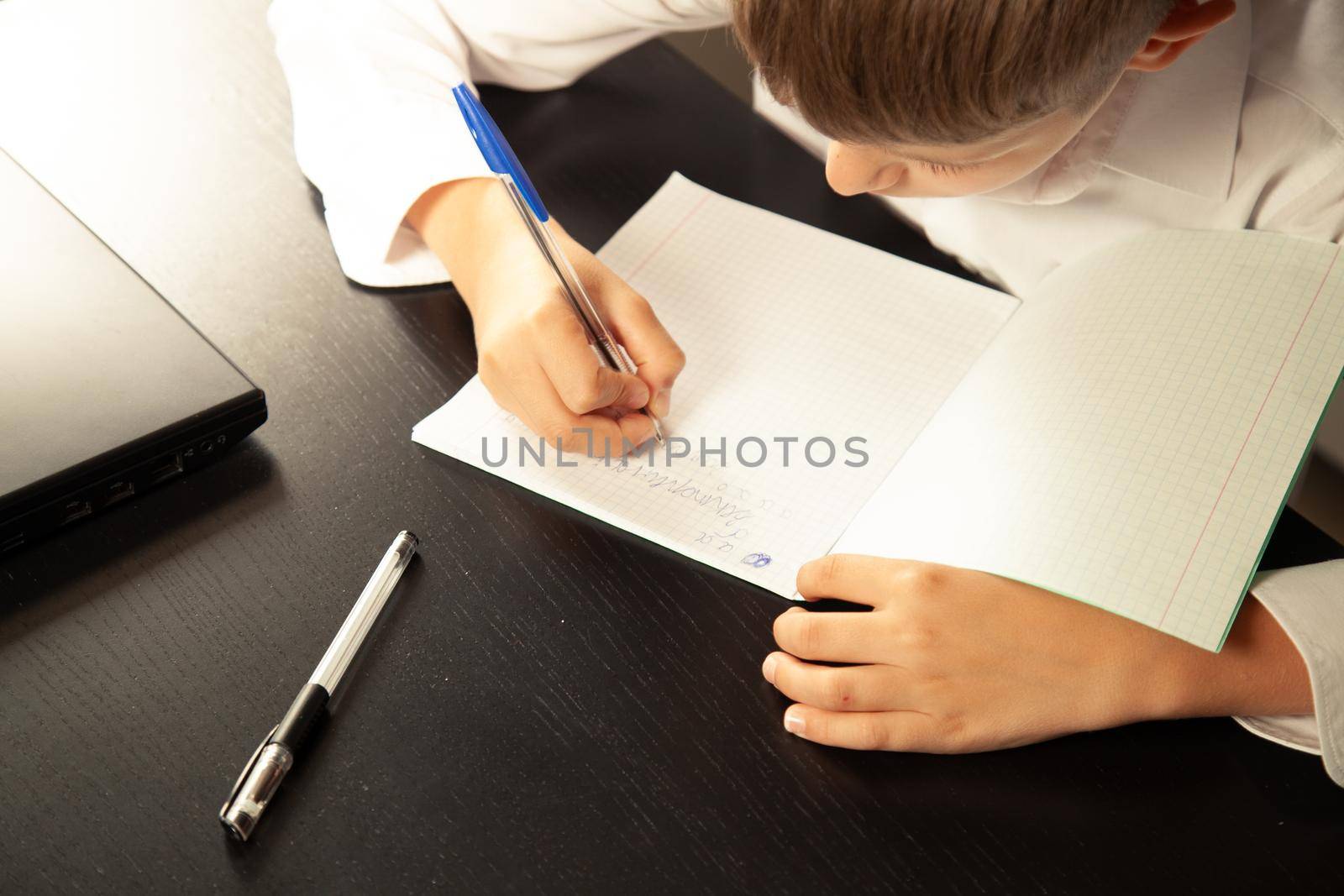 Close-up of a child writes in a notebook. Laptop and pencil on the table. Back to school