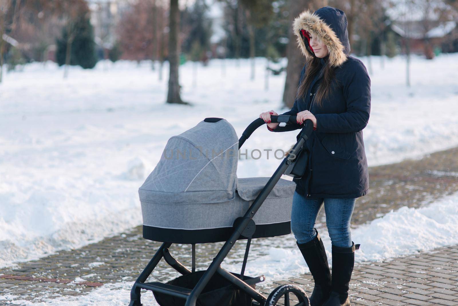 Beautiful mother walking in the park with her little baby in stoller. Woman dressed in blue jaket with hood and jeans. Warm boots.