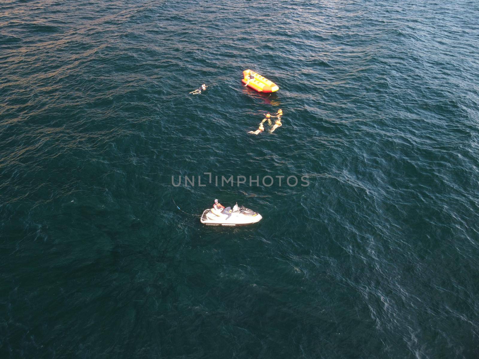 Happy people swim on air mattress behind a high speed water bike. Tourists ride on inflatable watercraft boat. Sea attraction. Water boat skating people on large Inflatable raft. Aerial copter view