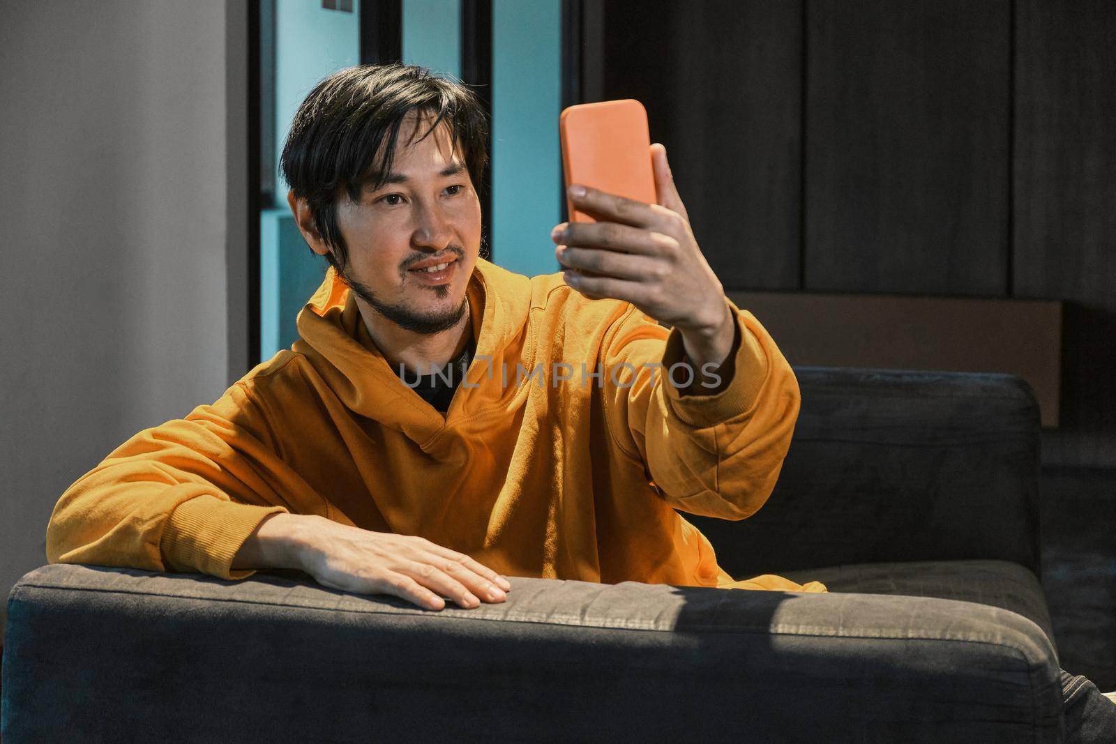 An Asian guy sits on sofa in a small office and communicates by mobile phone by snep_photo