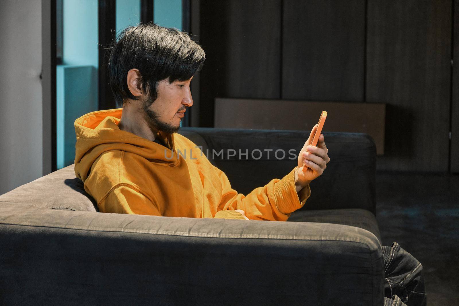 An Asian guy sits on sofa in a small office and communicates by mobile phone by snep_photo