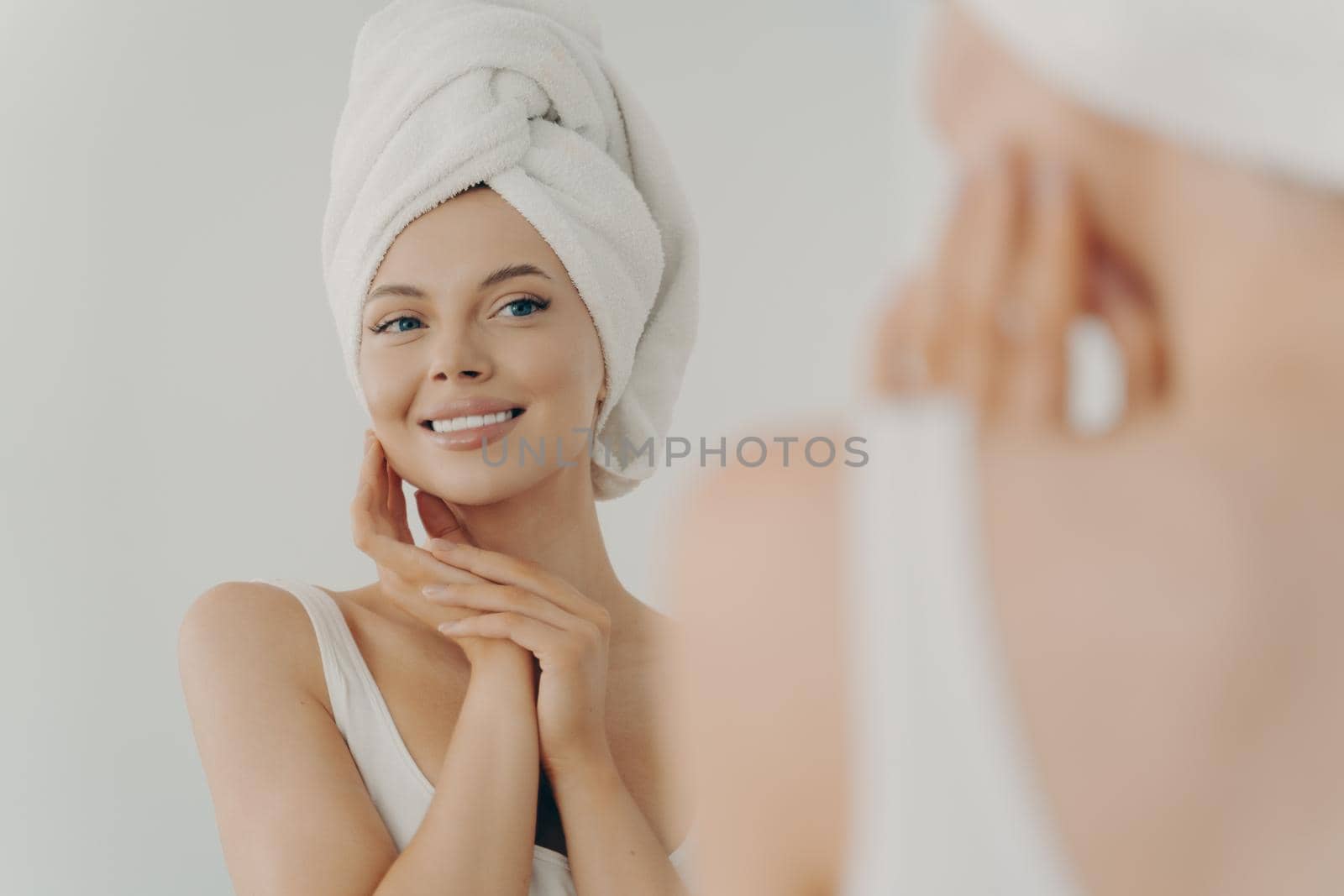 Beautiful young woman after shower looks in mirror gently touching soft healthy glowing face skin, happy millennial female applying beauty products during daily facial procedures, skincare concept
