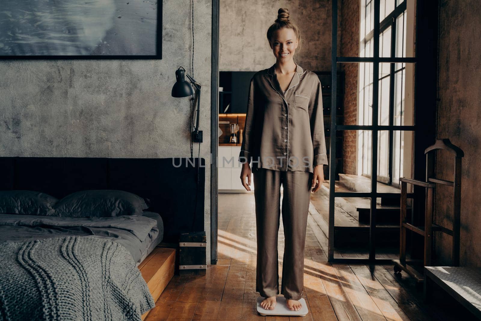 Dieting and weight loss. Full length of happy attractive young girl with hair tied up in bun in casual satin pajama standing on scales in stylish bedroom background and broadly smiling on camera
